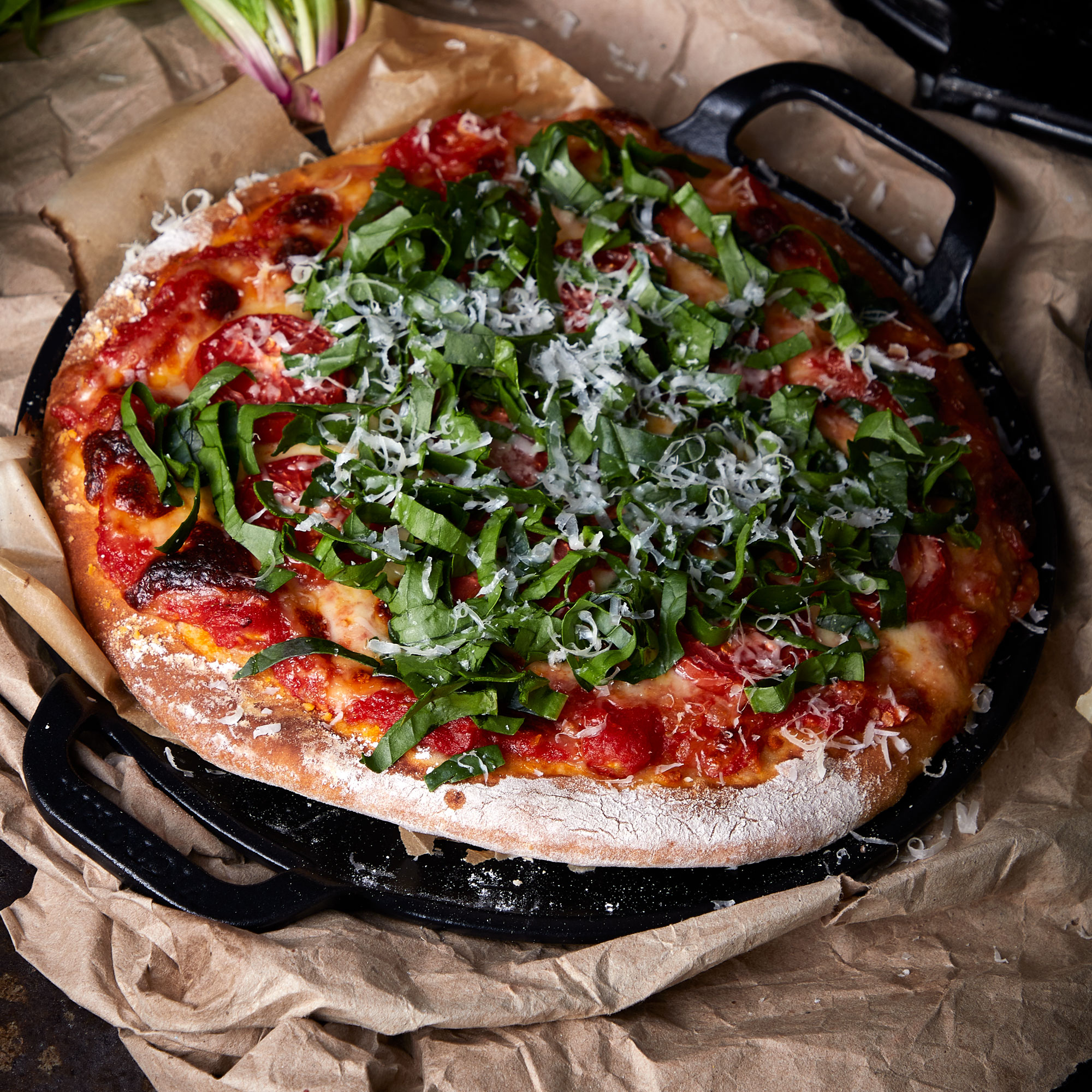 Pizza in einer Gusseisenpfanne, belegt mit Basilikum und Parmesan. Braunes Papier darunter.