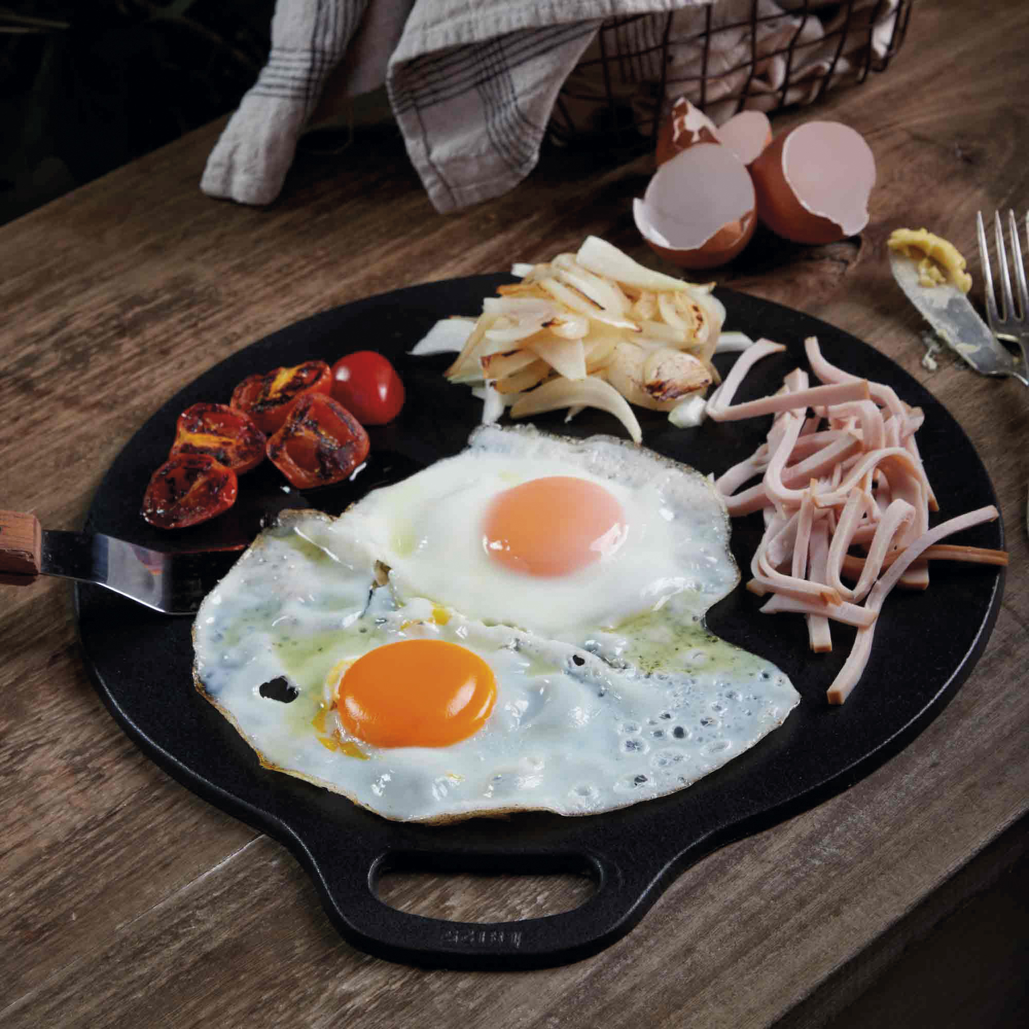 Spiegeleier, Tomaten, Zwiebeln und Schinken auf einer schwarzen Grillplatte, Holztisch, Gabel.