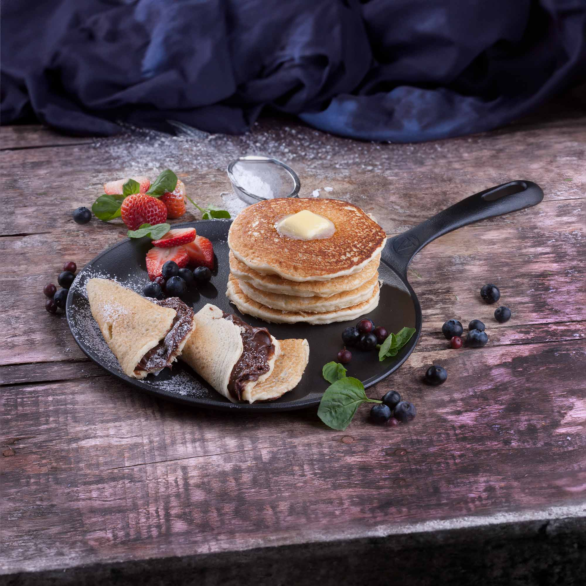 Pfannkuchen mit Butter und Beeren auf einer Pfanne. Crêpes, Erdbeeren und Blaubeeren sind auch auf dem Teller.