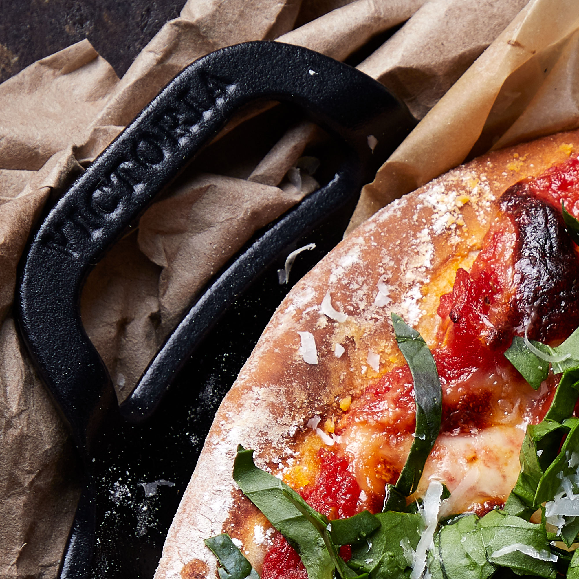Nahaufnahme Pizza in braunem Papier, schwarzer Pizzaschneider. Rote Soße, Käse, Basilikum.