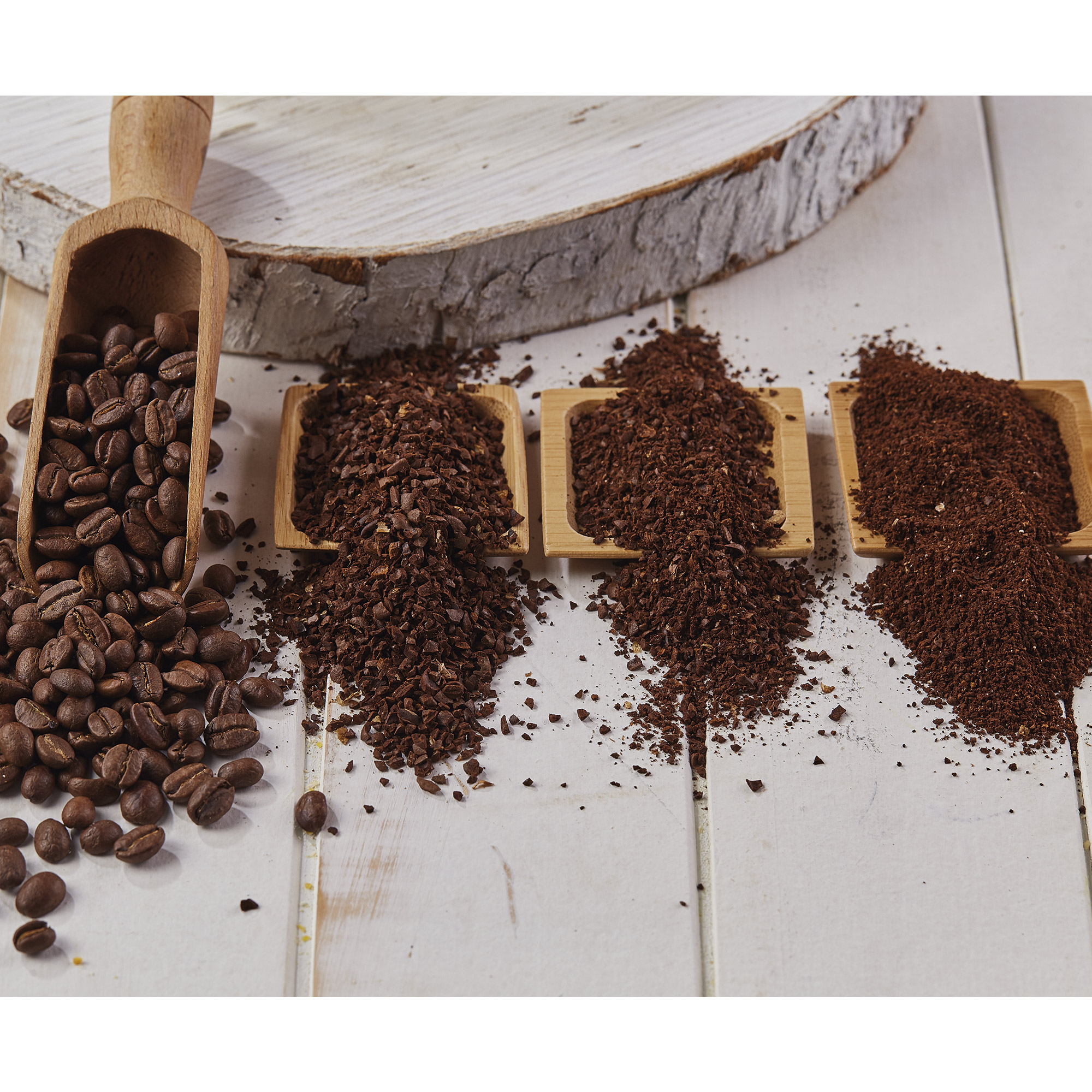 Granos y molido de café en cucharas de madera sobre una superficie de madera blanca. Diferentes molidos visibles.