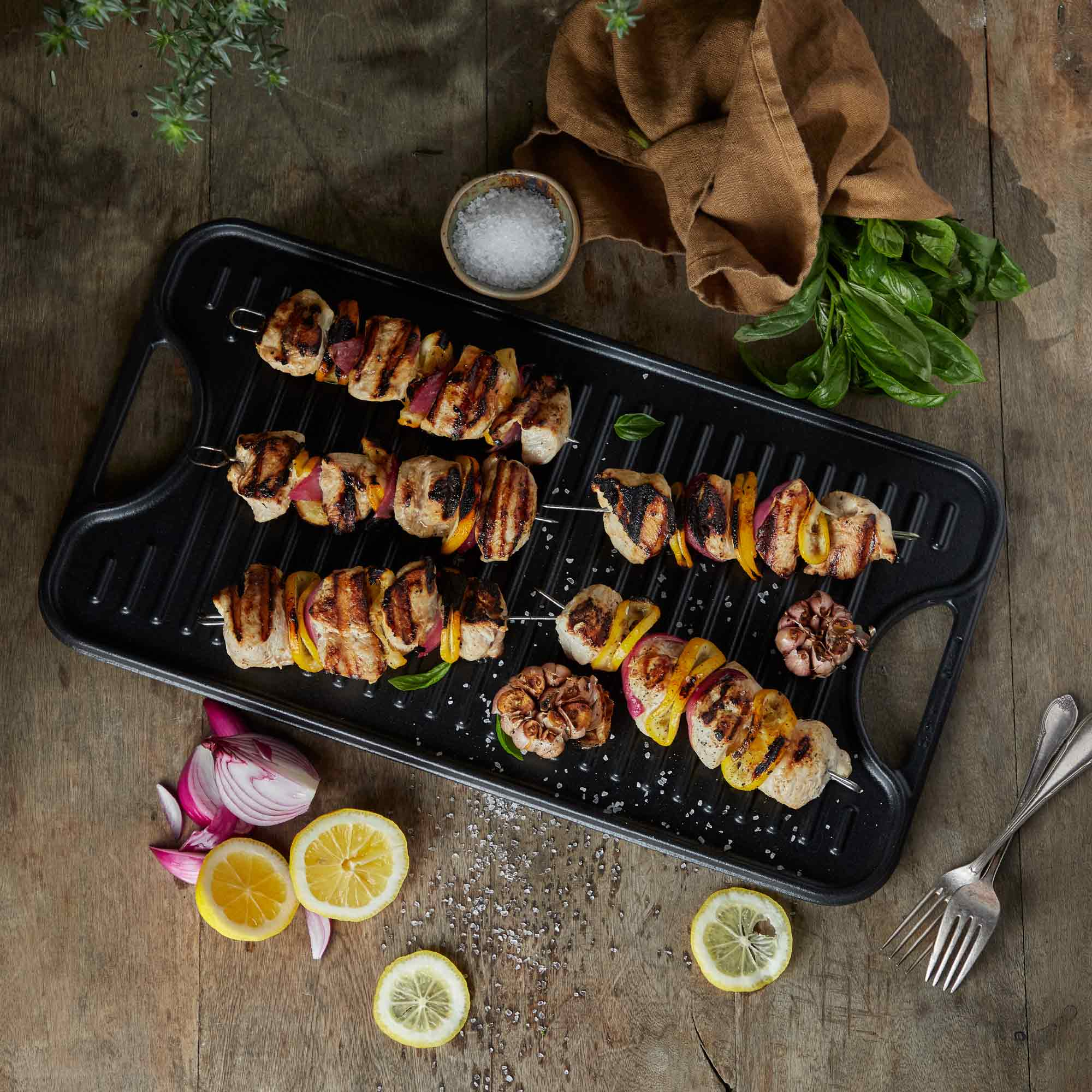 Gegrillte Spieße mit Fleisch, Zwiebeln und Paprika auf einer schwarzen Grillplatte. Frische Kräuter und Zitronen umgeben.