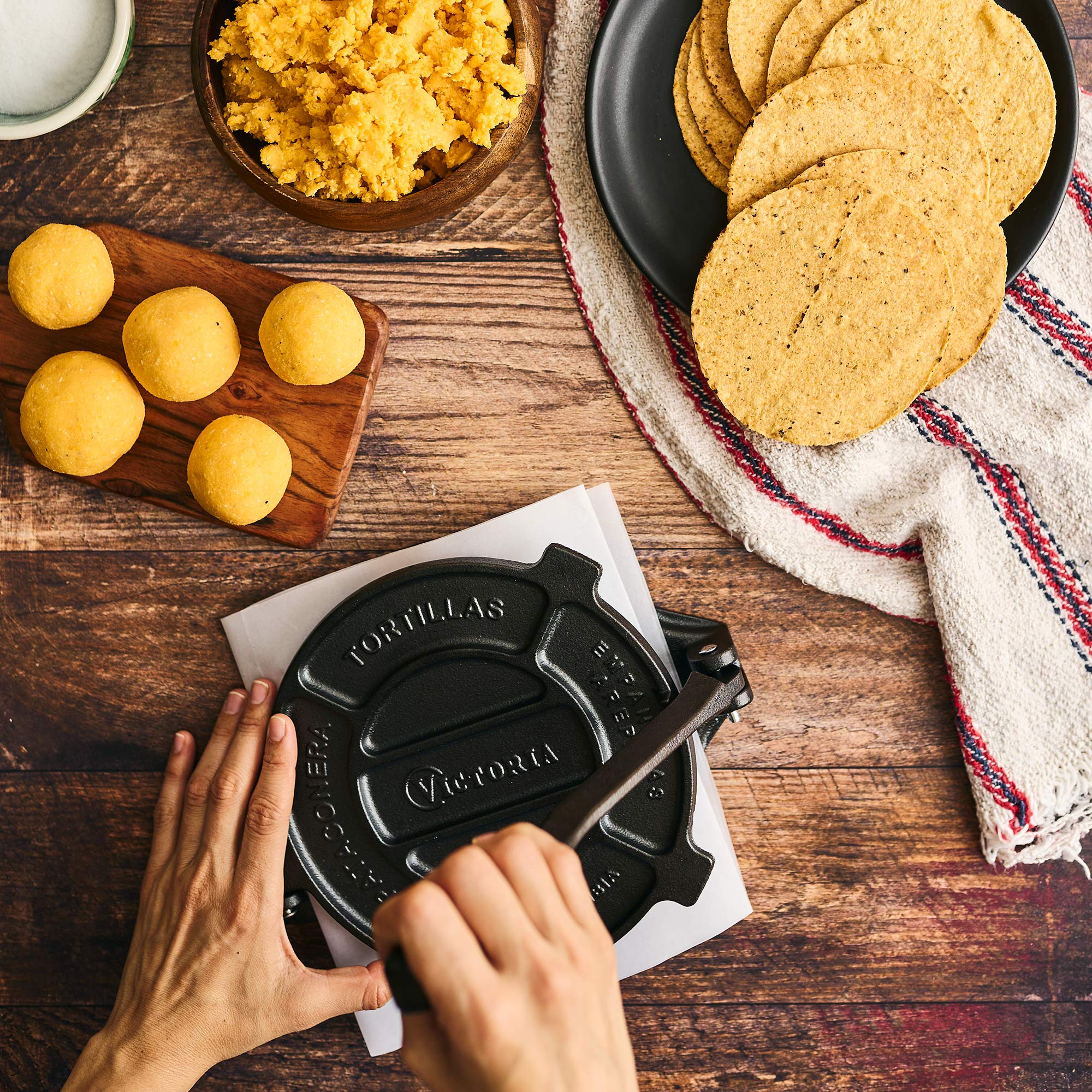 Draufsicht: Hände pressen Tortillas auf einer Tortillapresse, mit Zutaten auf einem Holztisch.