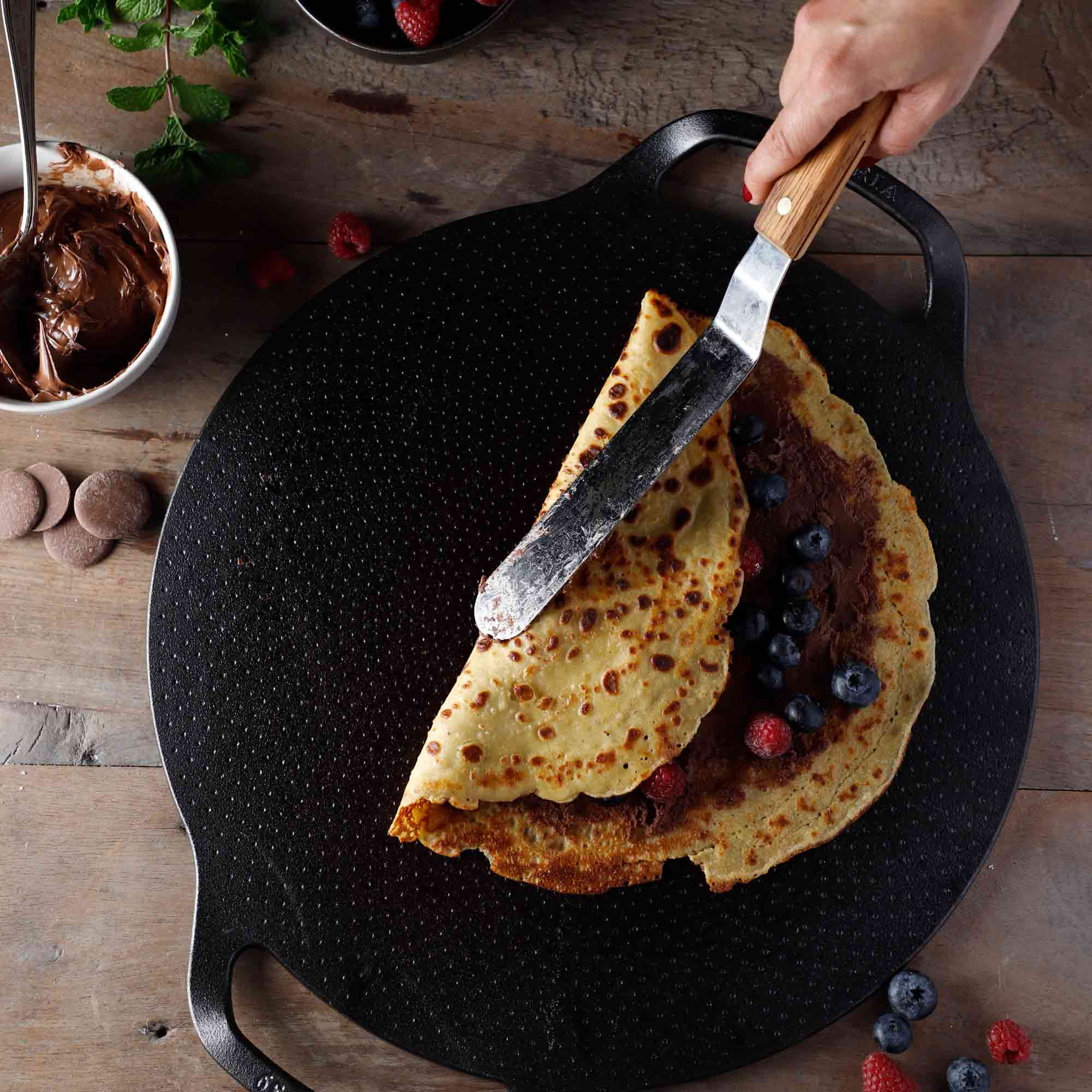 Pfannkuchen mit Schokoladenaufstrich, Blaubeeren und Himbeeren auf einer schwarzen Grillplatte zubereitet.