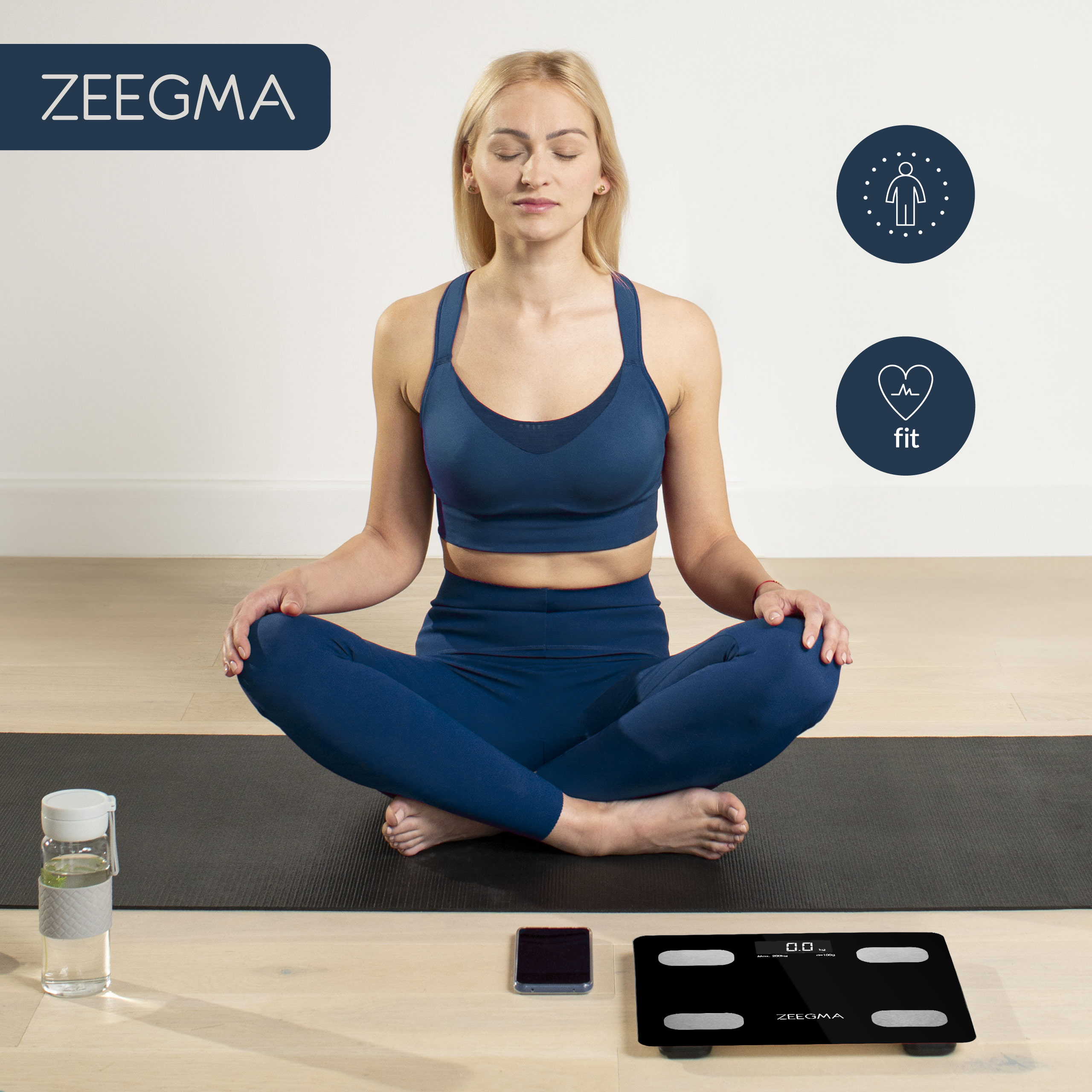 Kobieta medytuje na czarnej macie z logo ZEE GMA. Waga i telefon leżą na podłodze.
