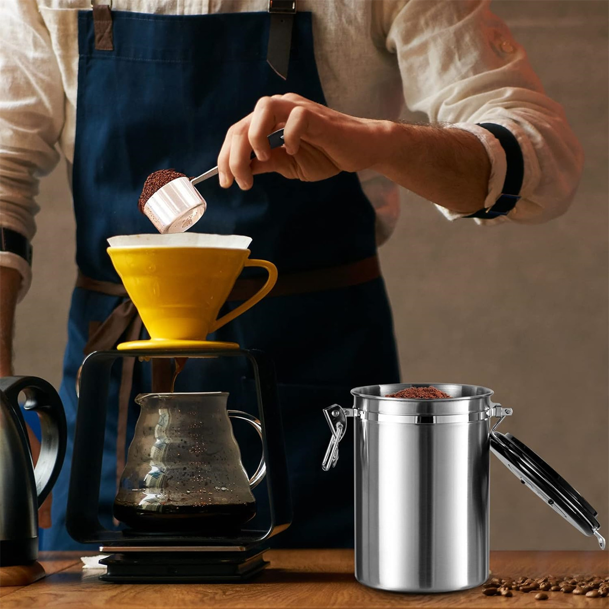 Eine Person gießt Kaffeepulver in einen gelben Pour-Over. Ein Metallkanister steht daneben.