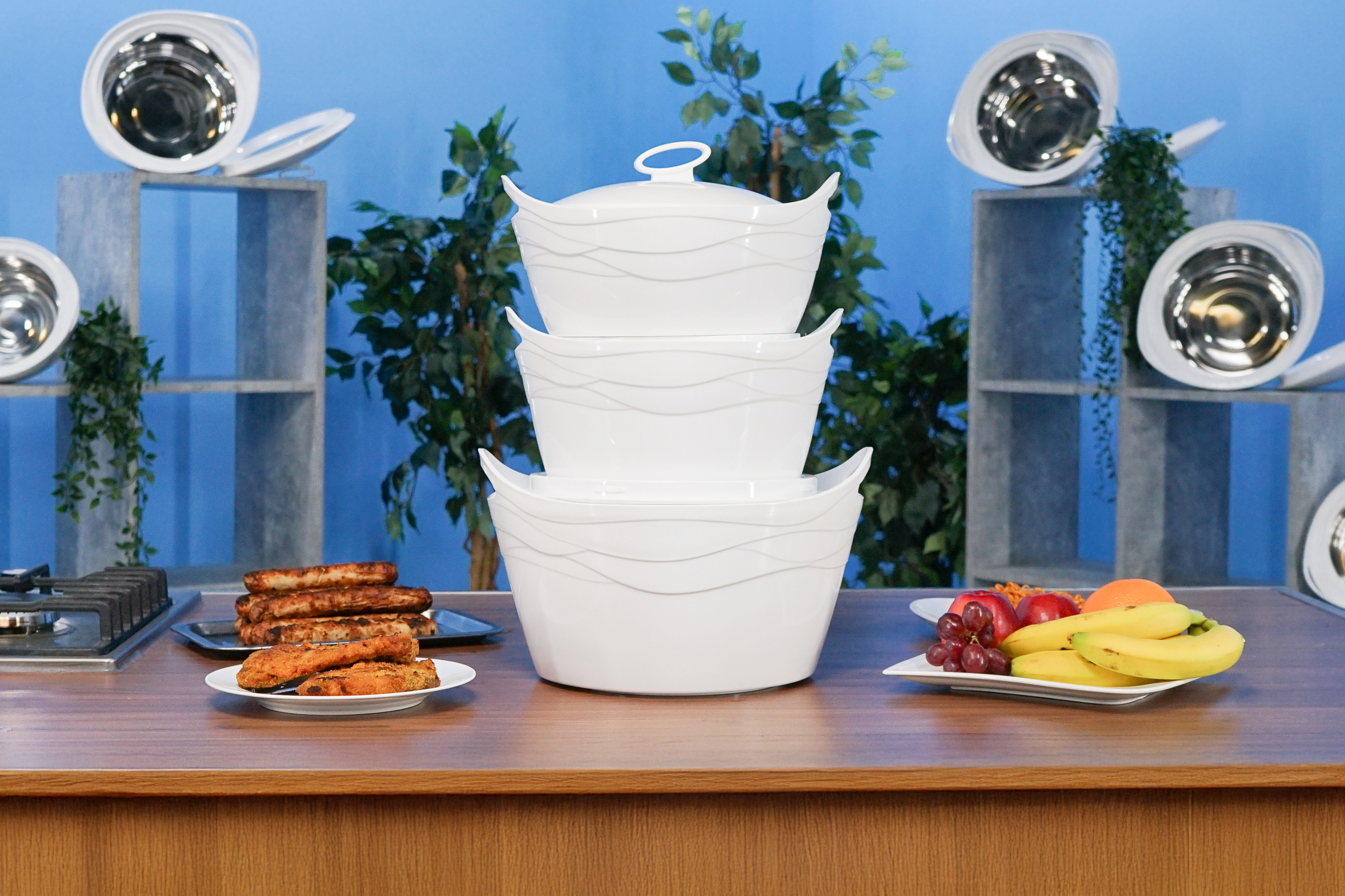 Tisch mit weißen Schalen, Tellern mit Essen und Obst. Hintergrund: blaue Wand und Regale mit Pflanzen.