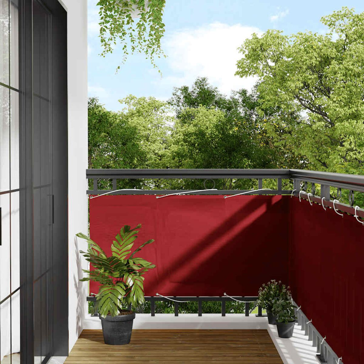 Balkon mit rotem Sichtschutz. Eine grüne Pflanze steht in einem Topf, im Hintergrund Bäume.