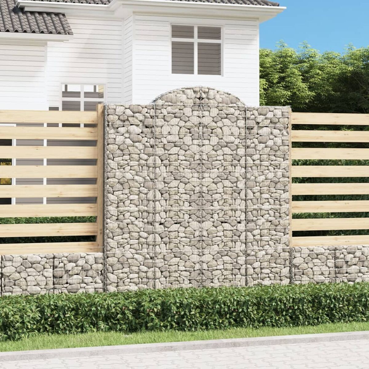Eine Steinmauer mit Holzzaun und Haus. Grüner Busch davor. Sonniger Tag.