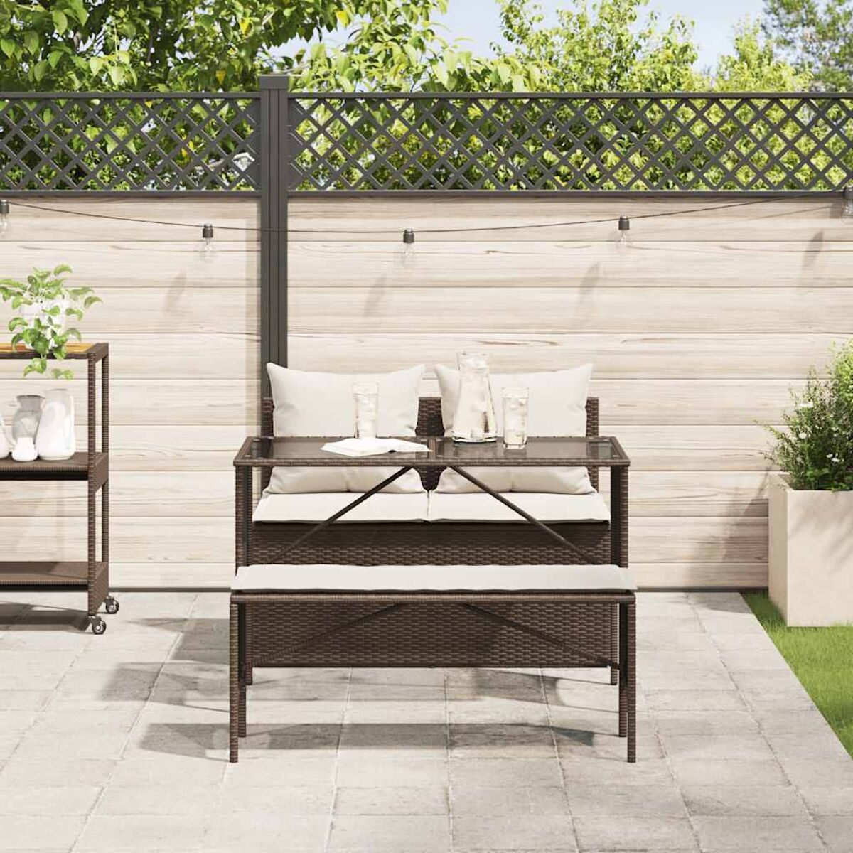 Ein braunes Rattan-Bank- und Tisch-Set mit Kissen, Lichtern und einem Holzzaun im Garten.