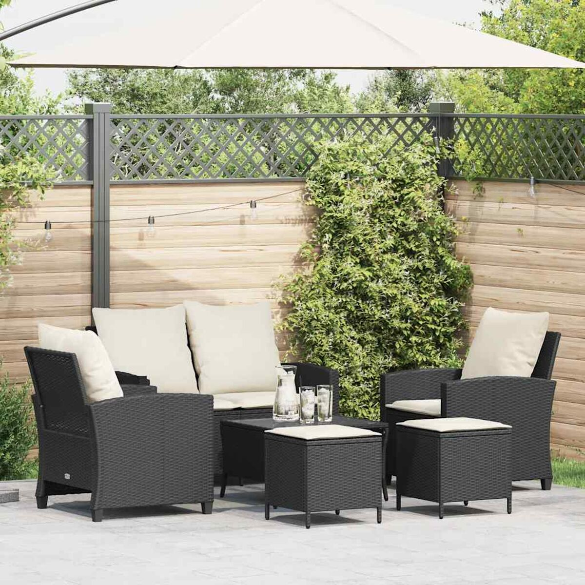 Set da esterno in vimini nero con cuscini beige, tavolo e ombrellone. Piante verdi e recinzione.
