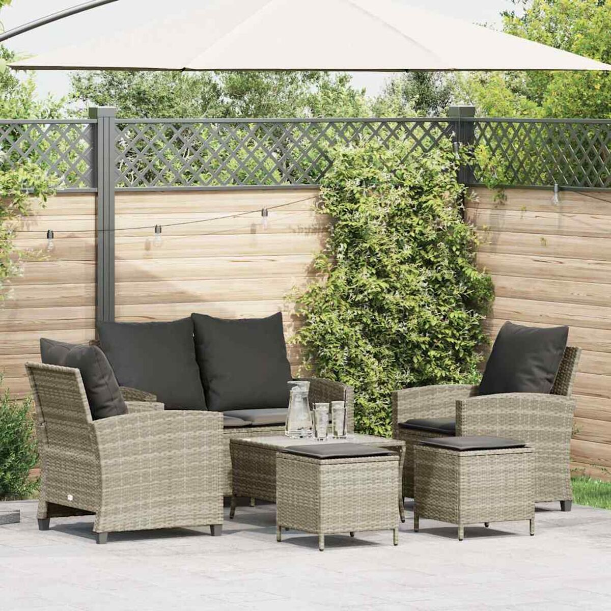 Set patio in vimini grigio con cuscini grigio scuro e un tavolo. Ombrello bianco e piante verdi.