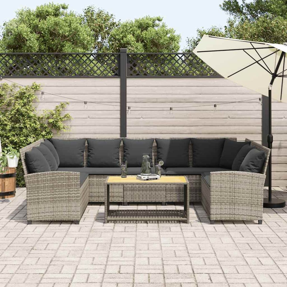 Terrassengarnitur: graues Korbmöbelsofa, schwarze Kissen, Holztisch und Sonnenschirm auf Steinterrasse.