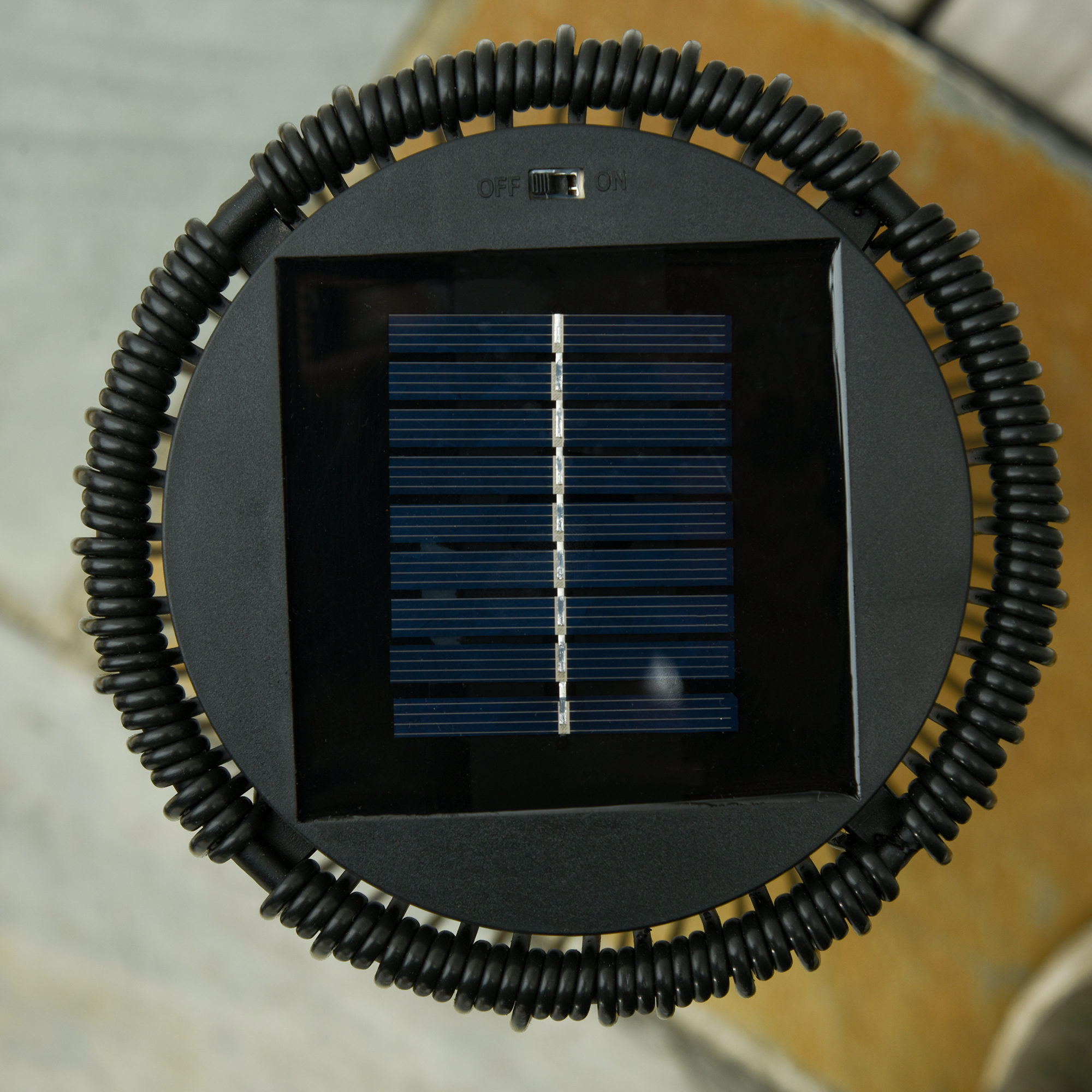 Schwarze Solar-Gartenleuchte mit Spulendesign und Solarpanel. Brauner Pflasterhintergrund.
