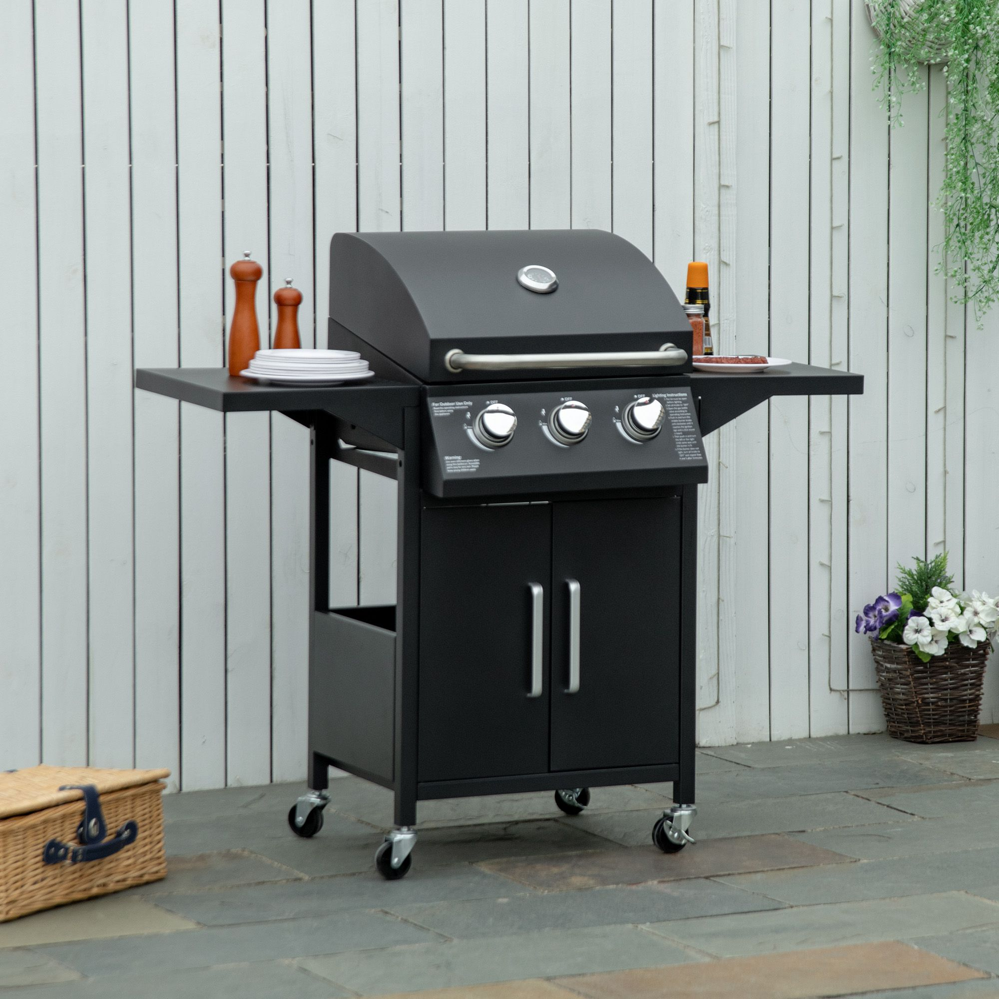 Ein schwarzer Gasgrill mit Seitentischen, Utensilien und einem Korb.