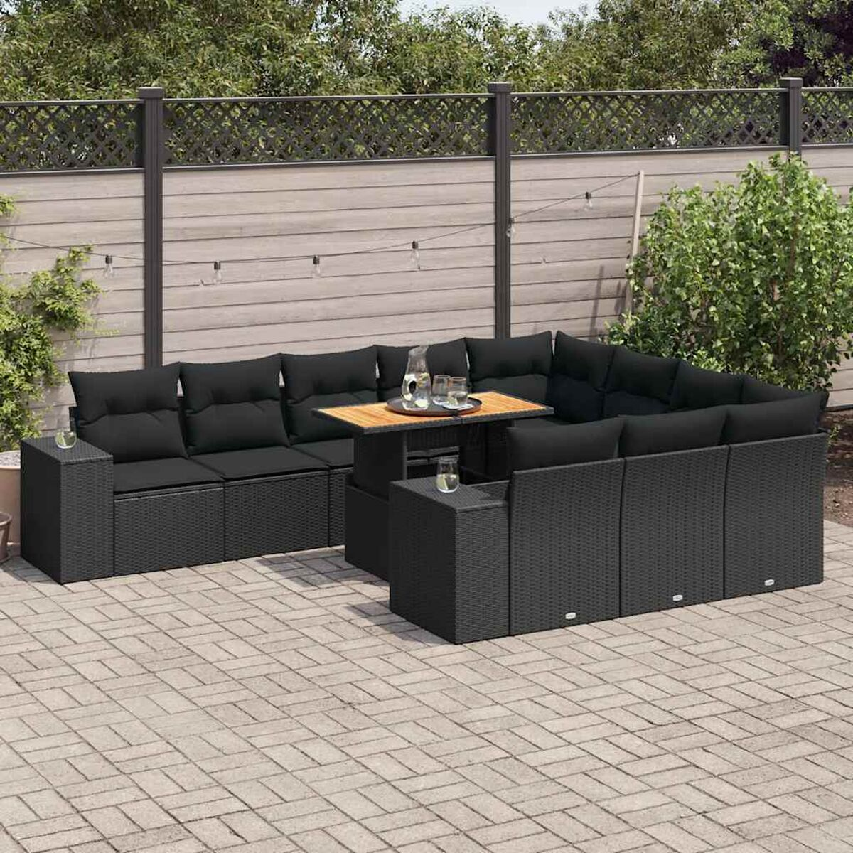Set patio all'aperto con cuscini neri, tavolo e contenitore. Una staccionata in legno e piante sullo sfondo.
