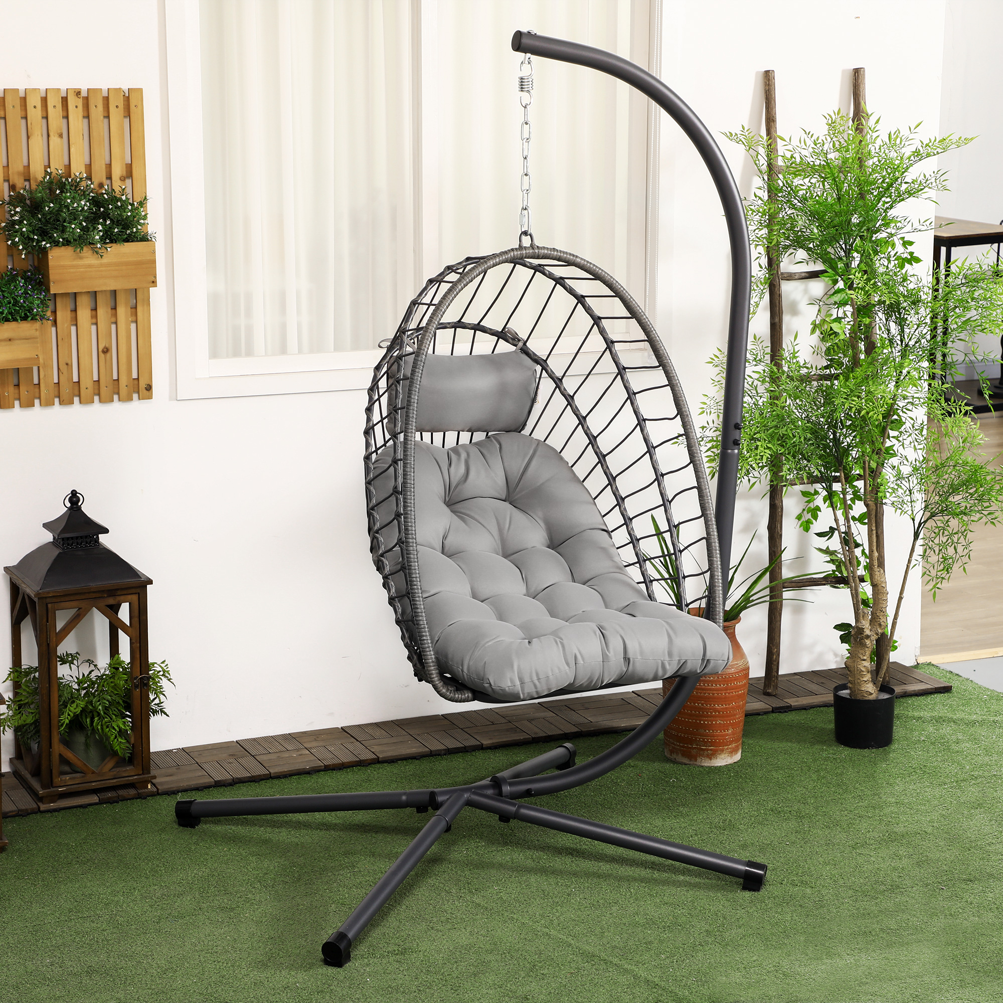 Silla colgante gris en un soporte de metal negro. En un patio trasero con hierba verde y plantas al fondo.