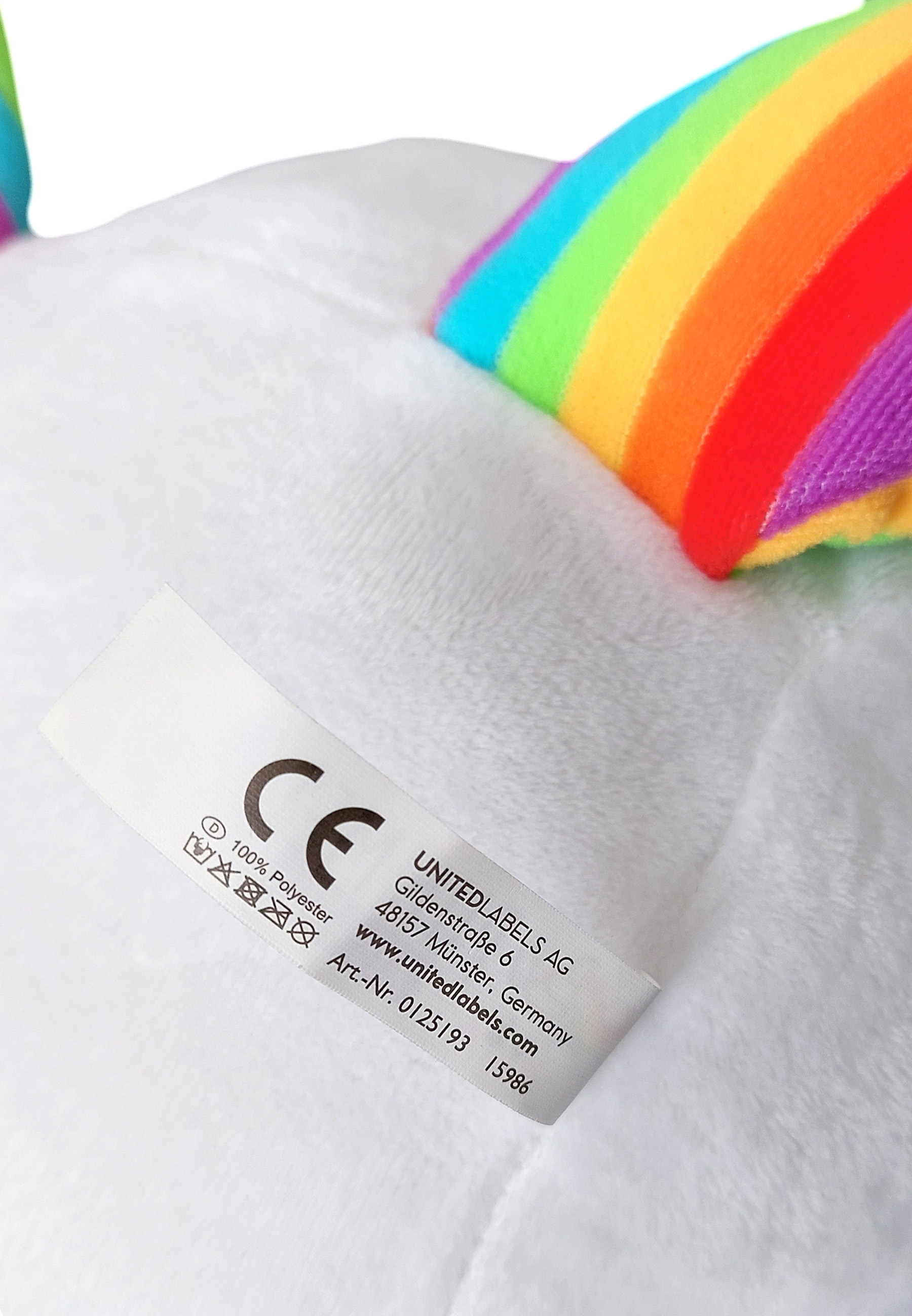 Ein weißes Stoff-Einhorn mit Regenbogenmähne und -schwanz. Ein Etikett mit Text ist am Einhorn befestigt.