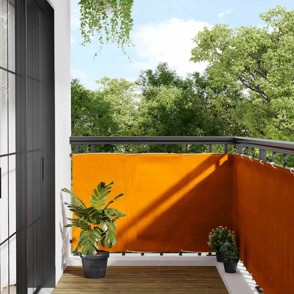 Balkon mit orangefarbener Sichtschutz, Pflanzen und schwarzem Geländer. Bäume und Himmel im Hintergrund.