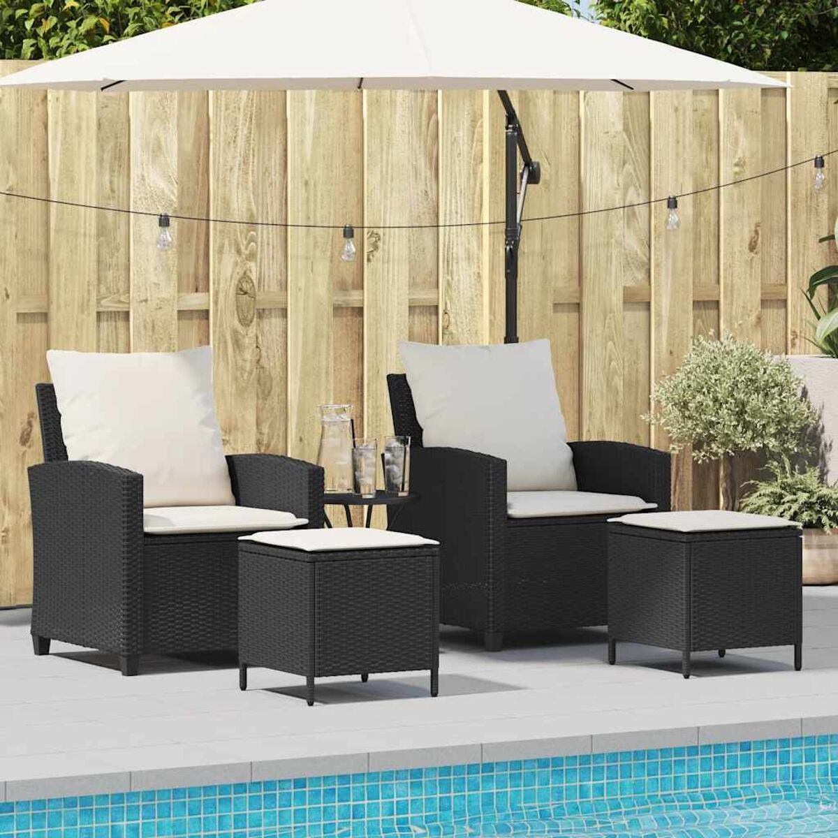 Area salotto all'aperto con due poltrone in vimini nere. Due pouf abbinati. Ombrellone bianco, piscina.