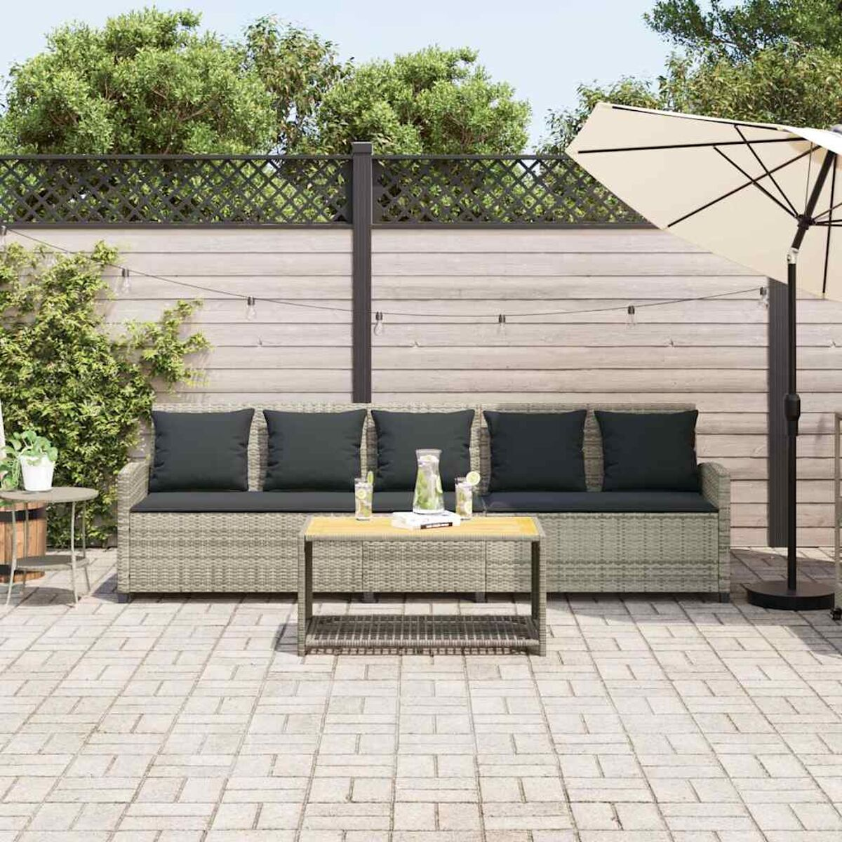 Allestimento patio con divano, tavolino e ombrellone. Pavimento lastricato, recinzione in legno e piante sullo sfondo.