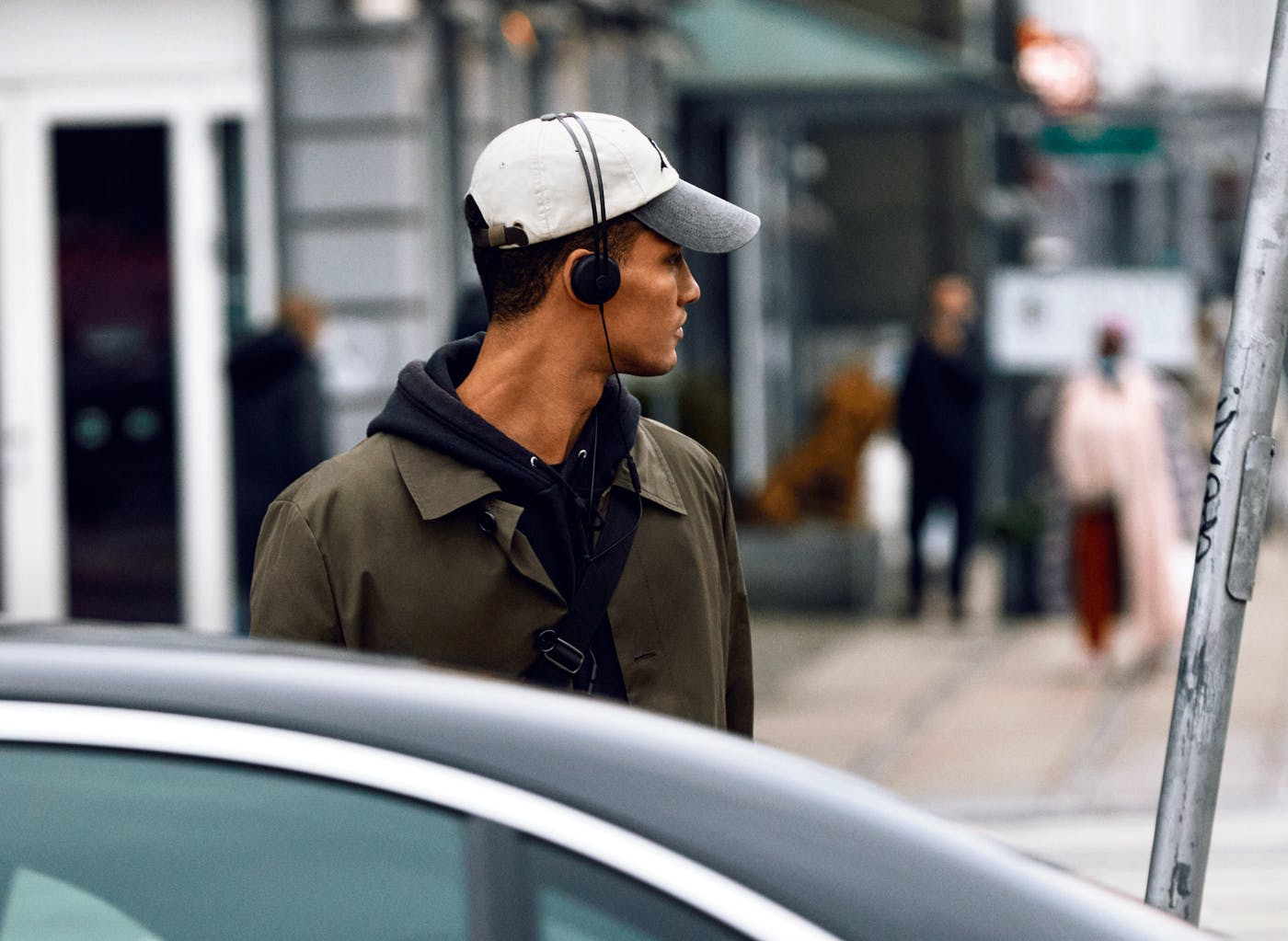 Uomo con cappello e cuffie, guarda altrove. Sfondo: edifici, persone, auto.