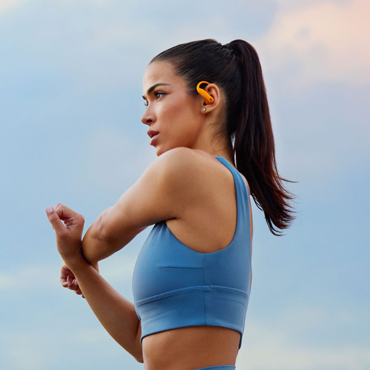 Kobieta rozciąga się na zewnątrz, w niebieskim topie i pomarańczowych słuchawkach. Tło nieba.