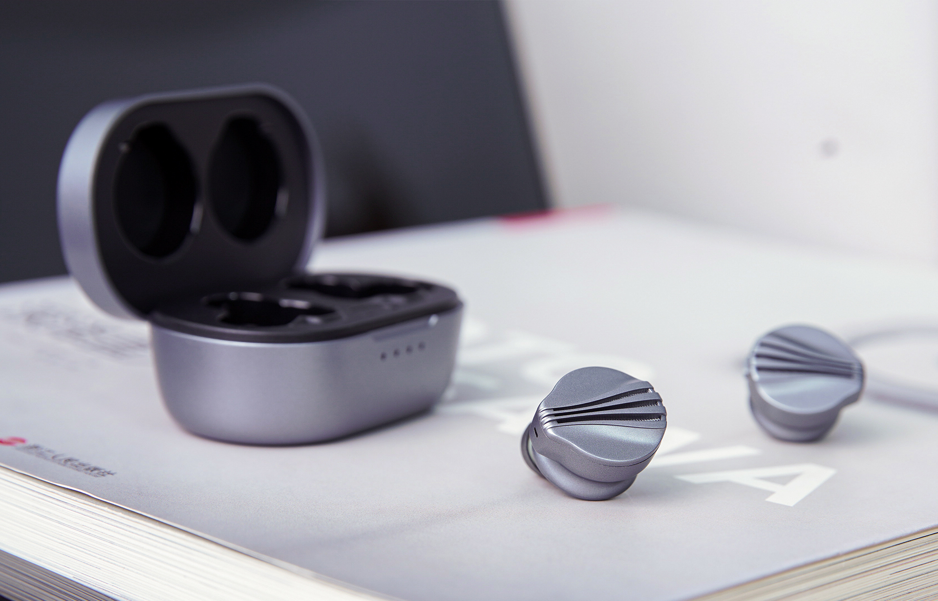 Twee grijze in-ear oortelefoons en oplaadcase, op een boek. Focus op de oortelefoons.
