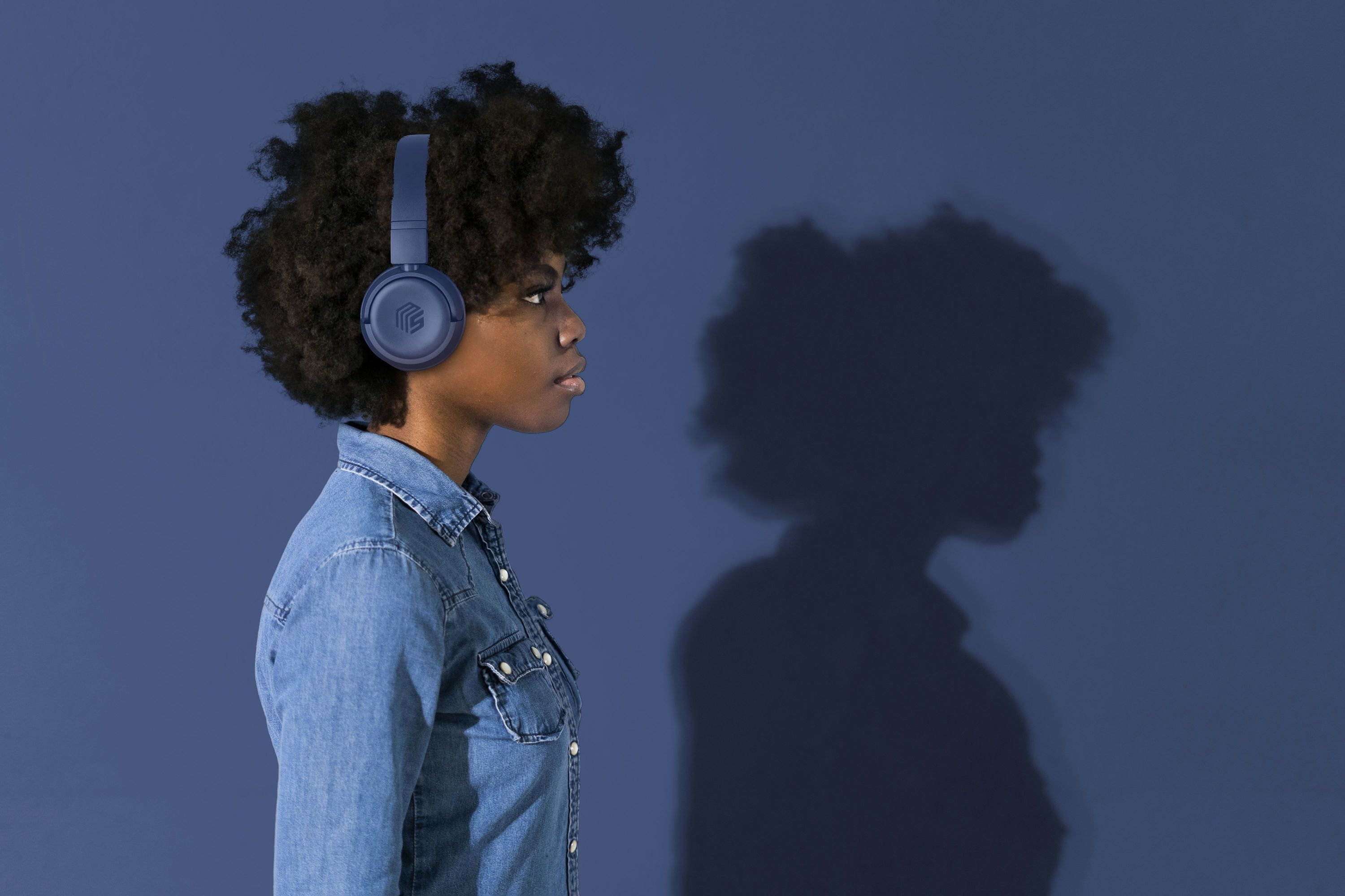 Eine Frau mit blauen Kopfhörern vor einer blauen Wand. Ihr Schatten ist an der Wand zu sehen.