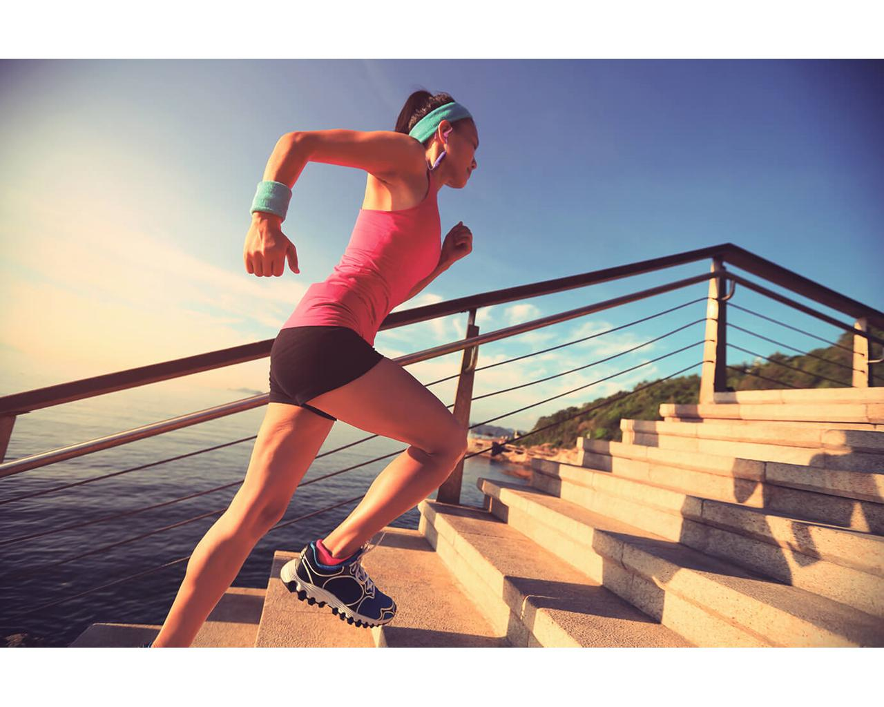Mujer corriendo escaleras cerca del océano, con ropa deportiva. Cielo azul en el fondo.