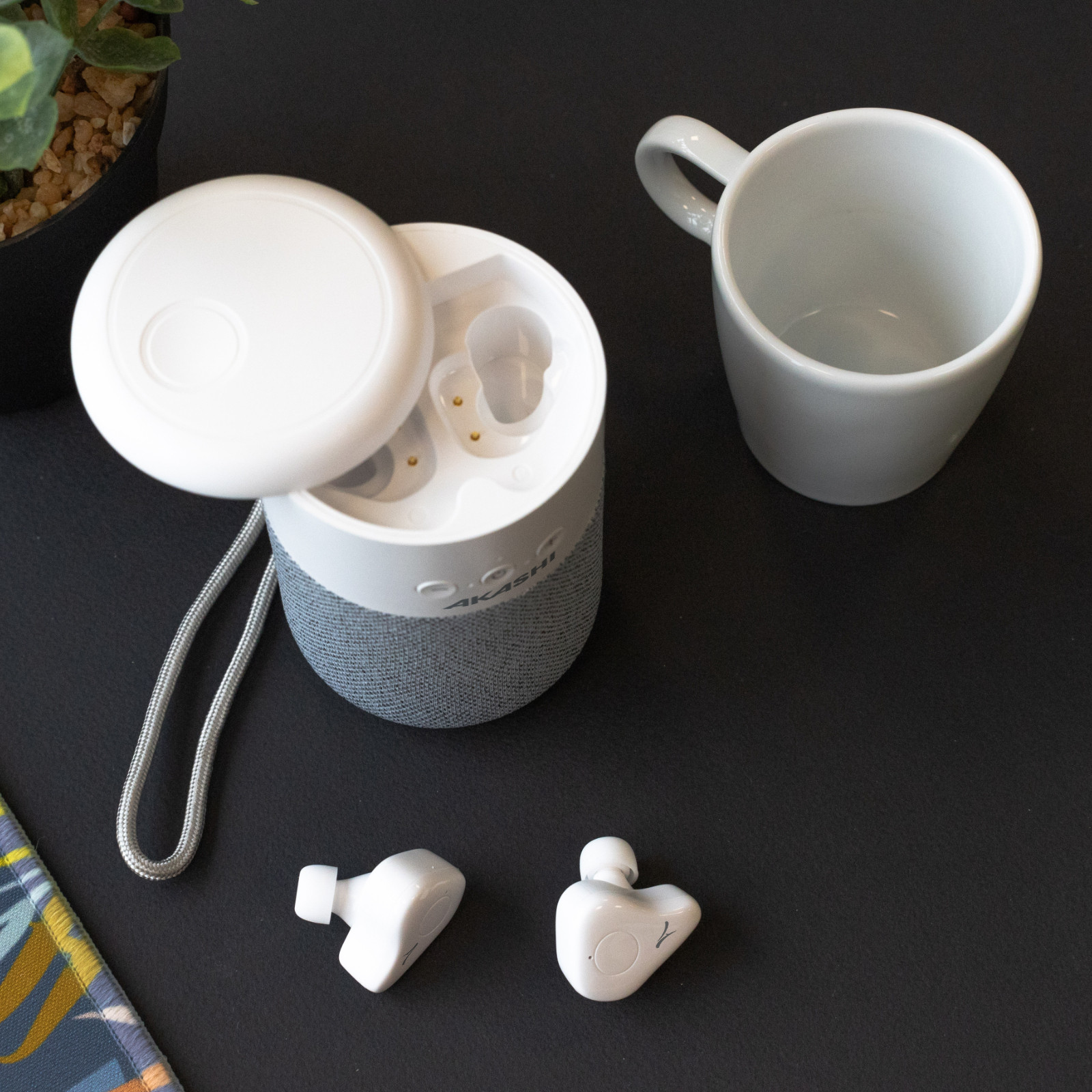 Weiße Ohrhörer, Ladecase und eine Tasse auf schwarzer Oberfläche mit Pflanze.