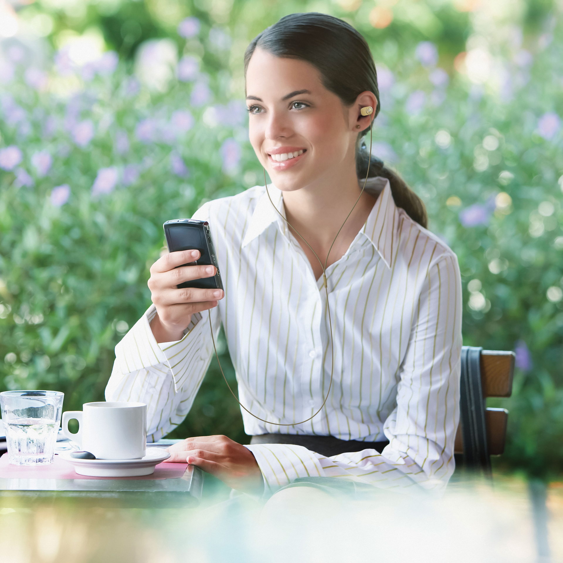 Uśmiechnięta kobieta, trzymająca telefon, w słuchawkach, na zewnątrz.