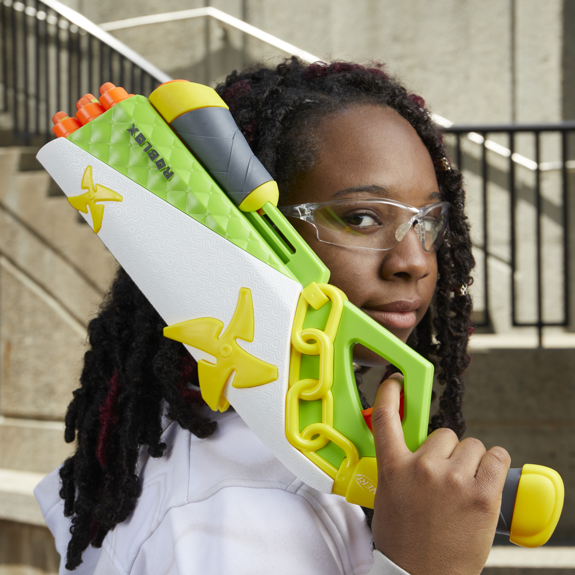 Una giovane donna tiene in mano una pistola giocattolo. La pistola è verde, bianca e arancione. Indossa occhiali protettivi.