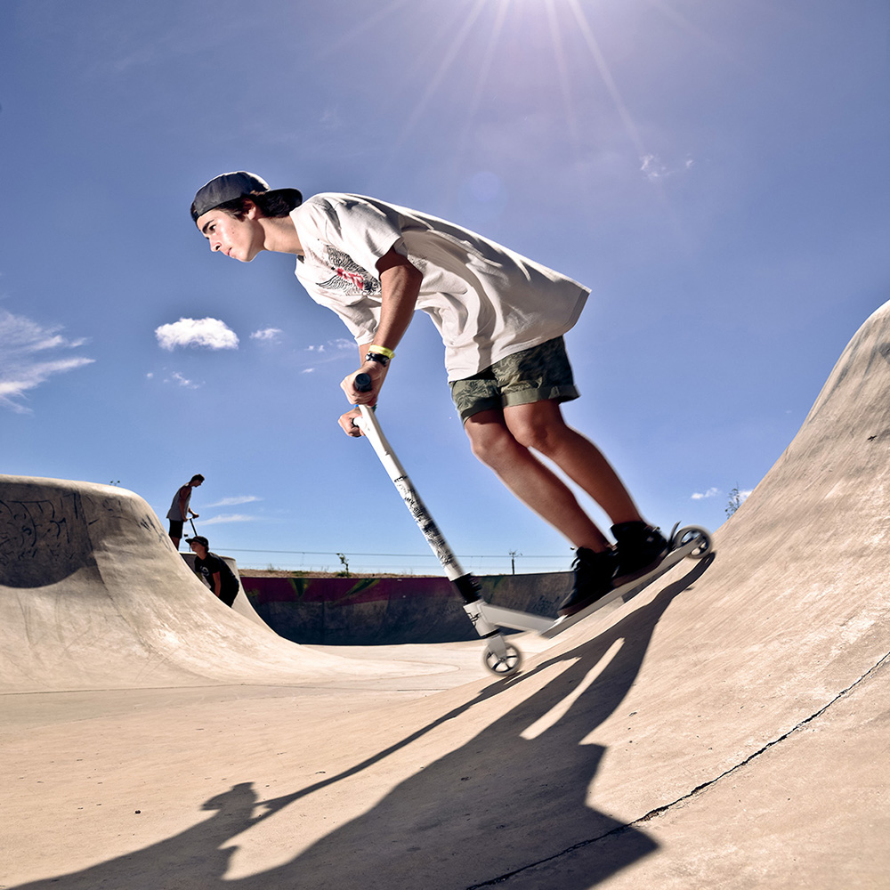 Giovane su uno scooter che esegue un trick in uno skate park. Cielo blu con nuvole. Rampa di cemento.