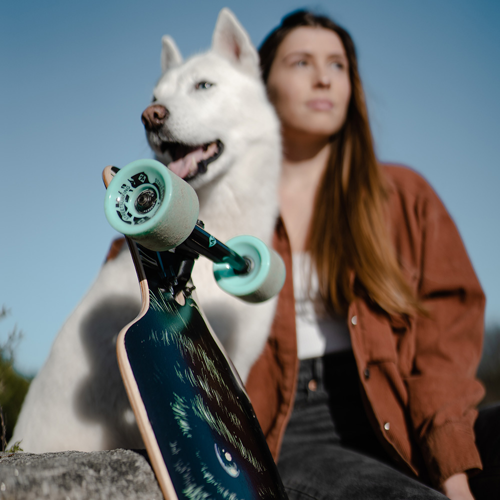 Una donna e un husky bianco con uno skateboard. Lo skateboard è a fuoco. La donna indossa una giacca marrone. Cielo sereno.