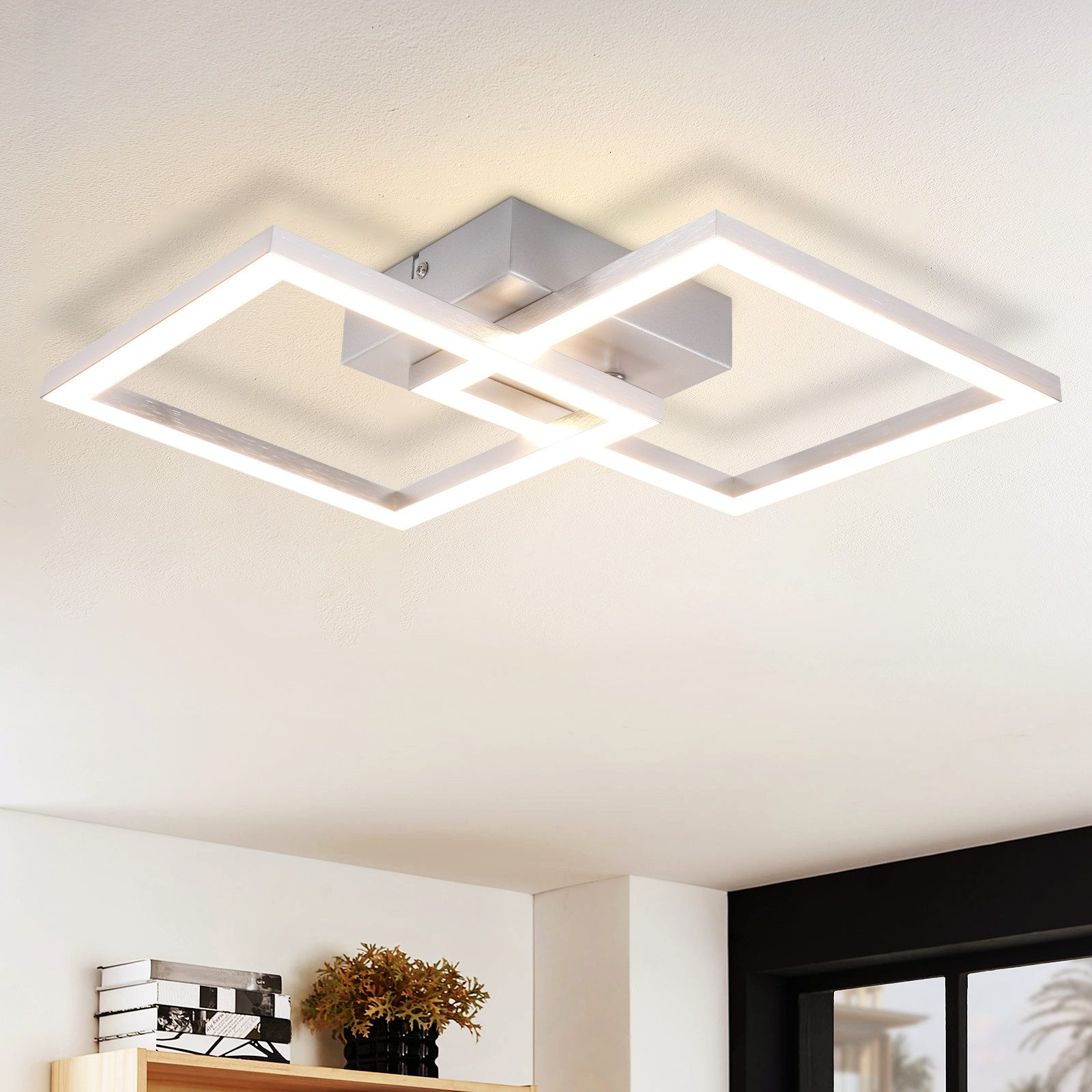 Nowoczesna lampa sufitowa z dwiema przecinającymi się kwadratowymi ramami, srebrną podstawą, na białym suficie.