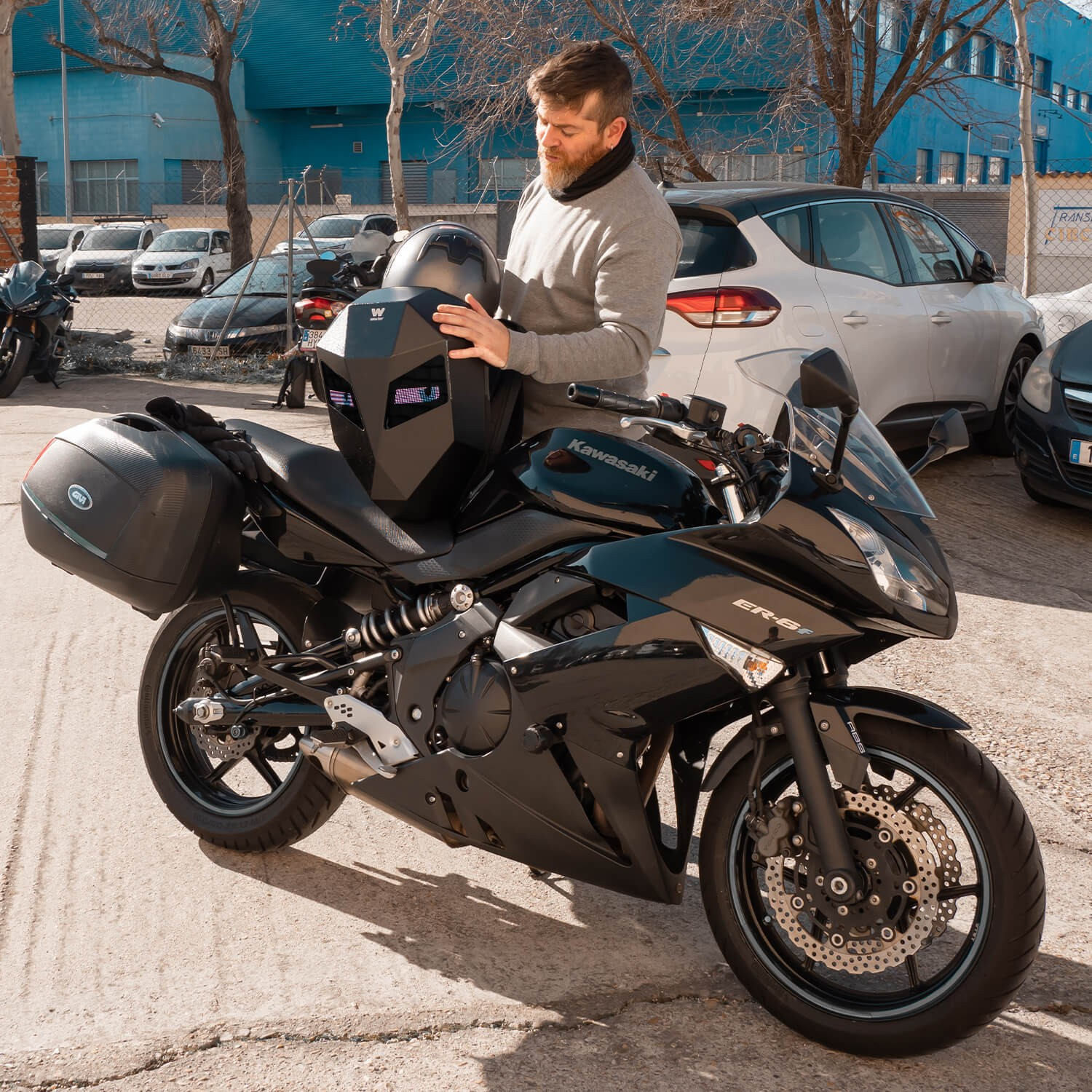 Hombre con casco junto a una moto negra aparcada, entorno exterior.