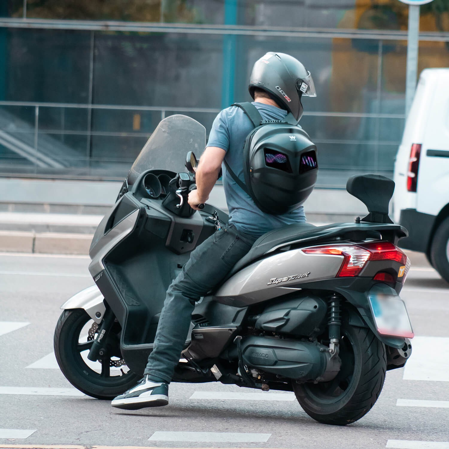 Persona montando scooter, con casco y mochila. Fondo borroso de carretera y edificios.