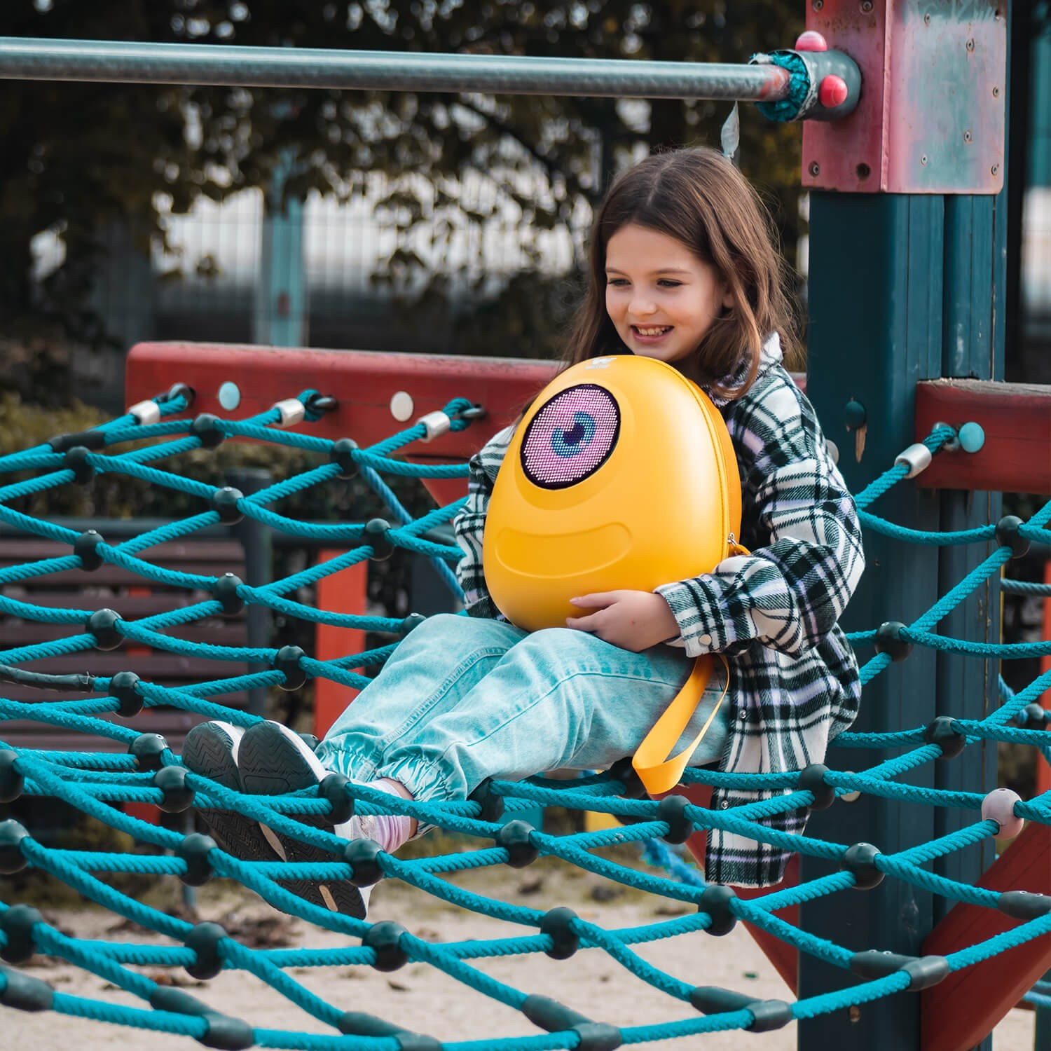 Una niña sentada en una red de juego, sosteniendo una mochila amarilla.
