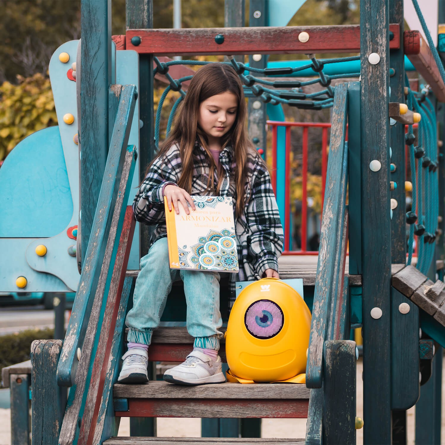 Una niña en un parque infantil leyendo un libro. Un robot amarillo a sus pies.
