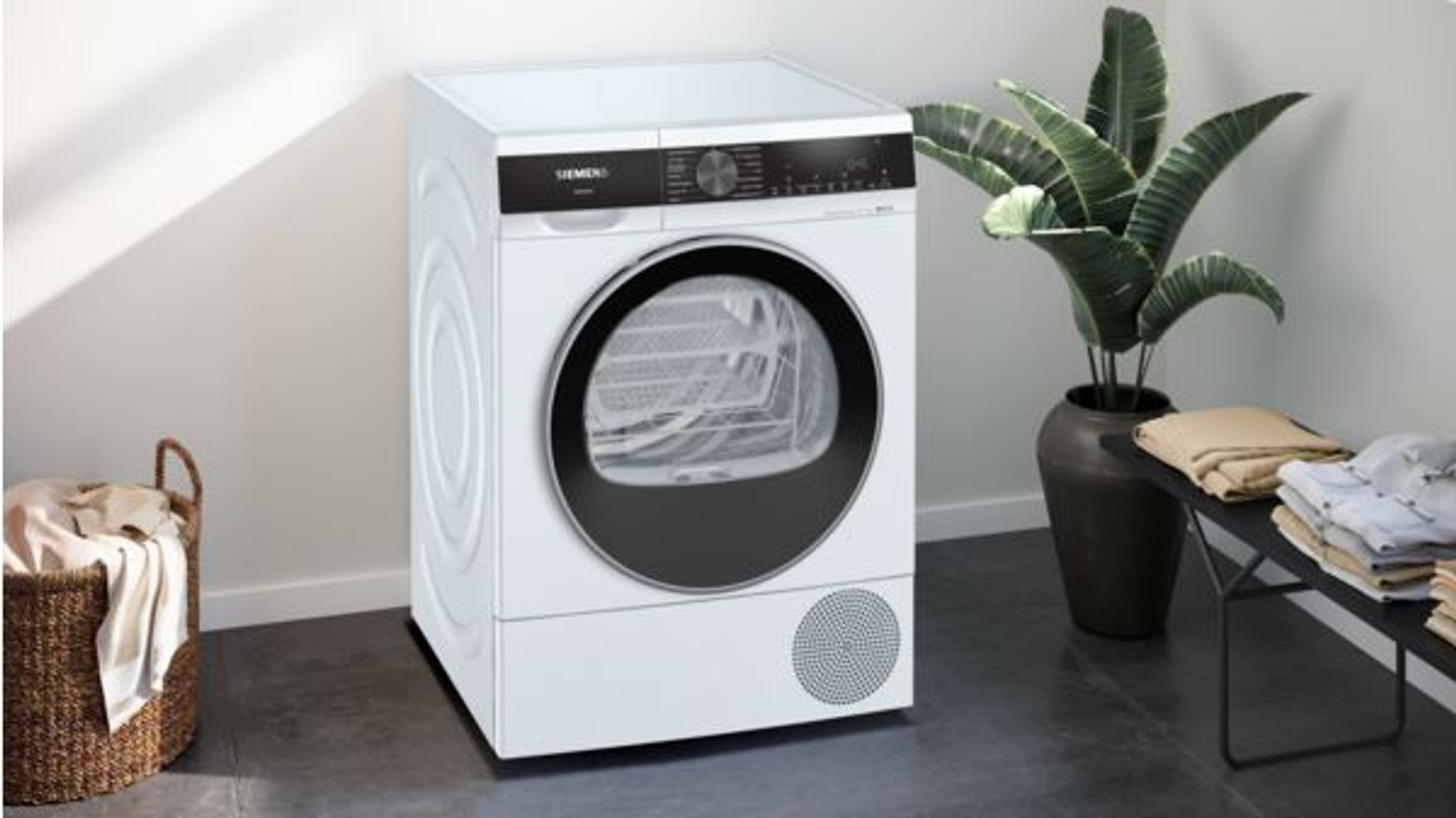 Secadora Siemens blanca en una habitación. Una cesta de ropa y una planta están a la izquierda y a la derecha.