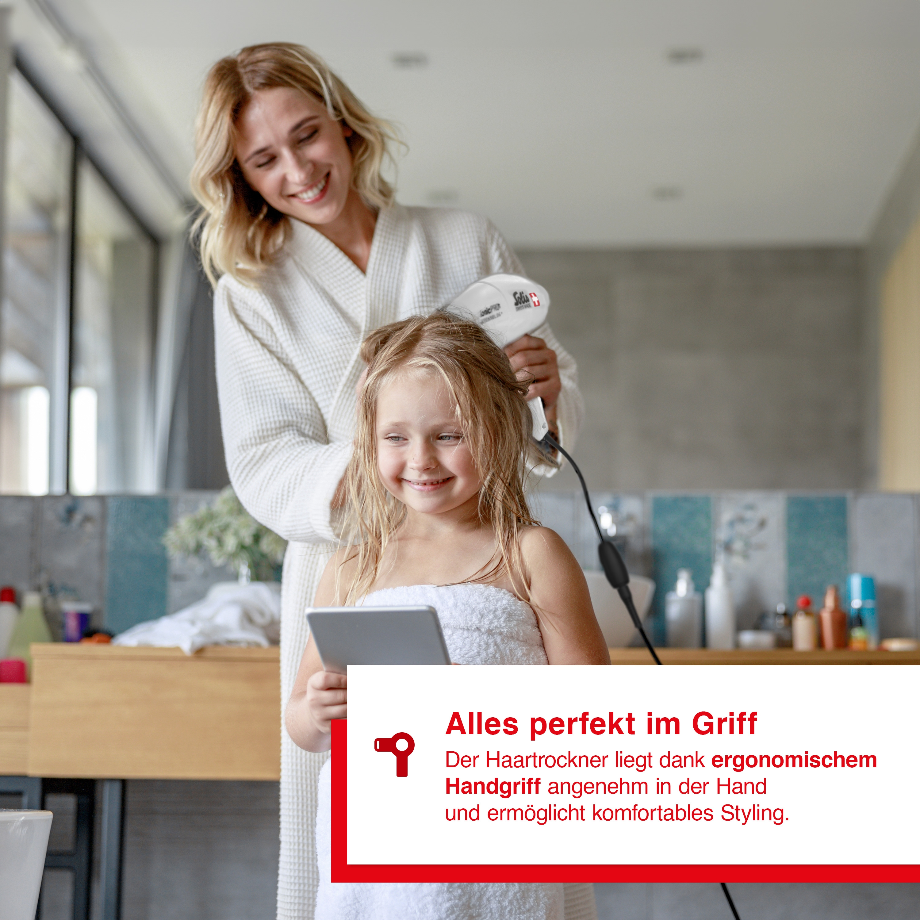Una donna asciuga i capelli di una bambina con un asciugacapelli in bagno, sorridendo.
