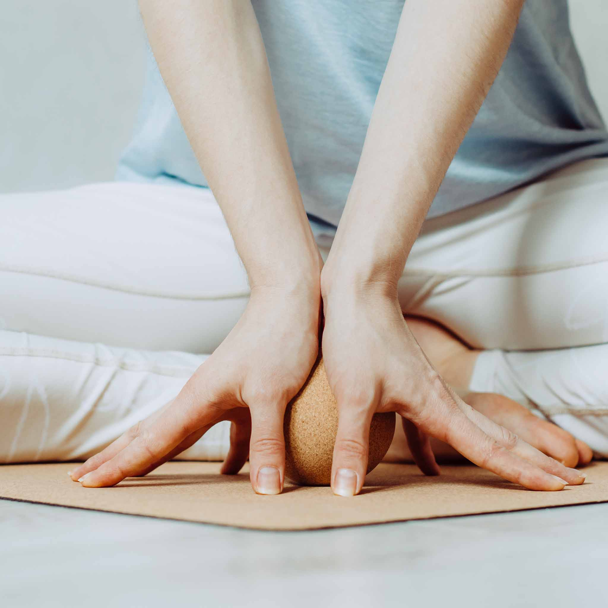 Person sitzt im Schneidersitz, Hände umschließen einen kleinen braunen Ball, auf einer Yogamatte.