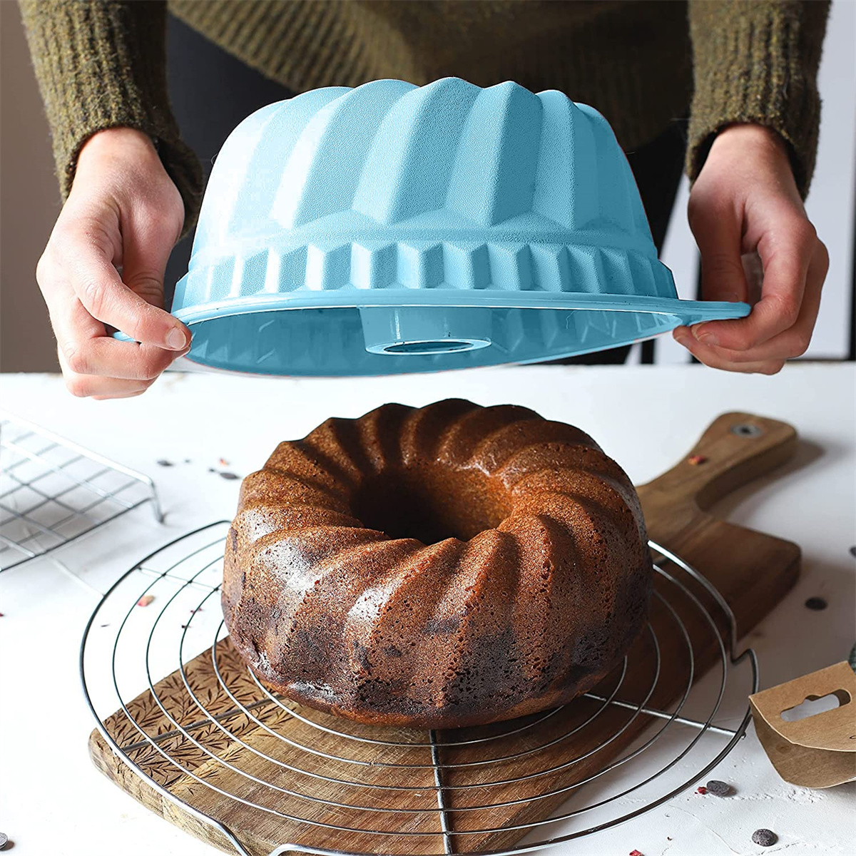 Frau hält eine blaue Gugelhupfform über einem Kuchen auf einem Kühlrost. Holzgrund.