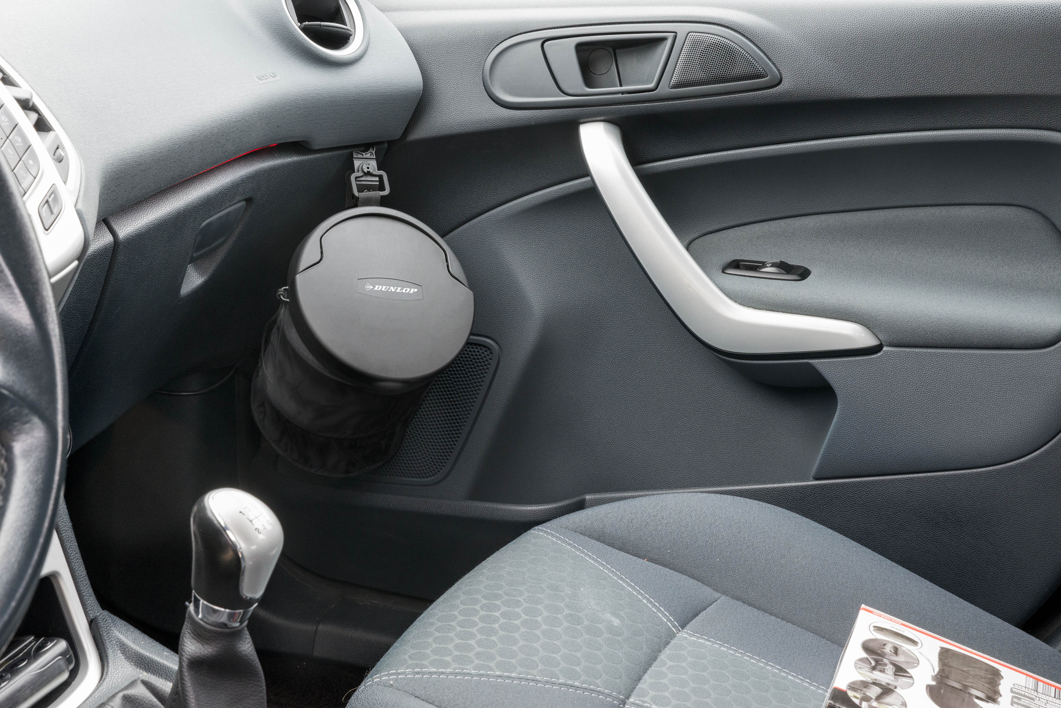 Auto-interieur met zwarte vuilnisbak aan de deur. Zilveren deurgreep en zwarte versnellingspook zichtbaar.