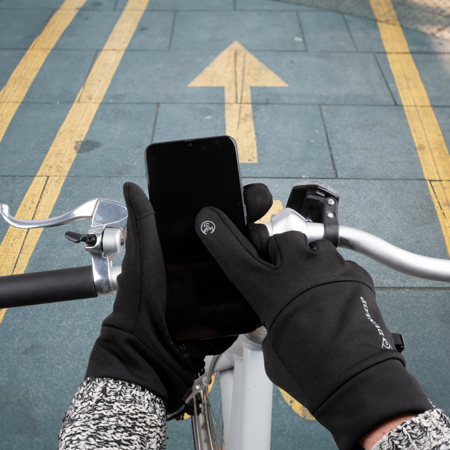 Handen in zwarte handschoenen houden een zwarte smartphone vast op een fietsstuur.