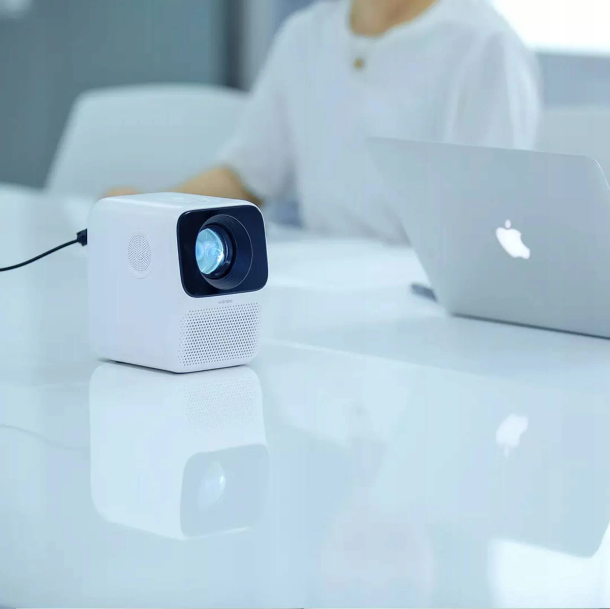 Proyector blanco sobre una mesa blanca, con un portátil al fondo.