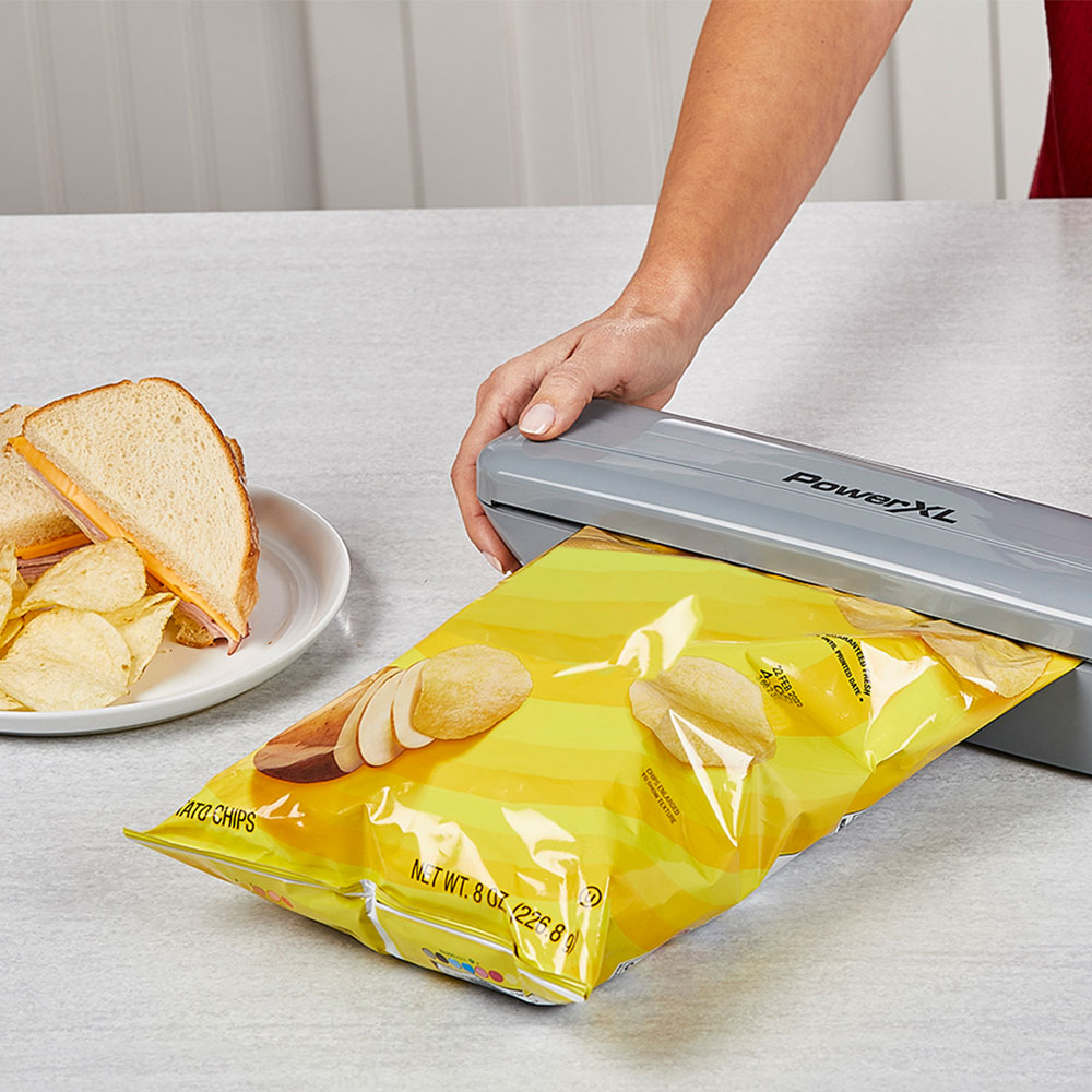 Eine Person verschließt eine Tüte Kartoffelchips mit einem grauen Heißsiegelgerät. Ein Teller mit Chips und Sandwich ist sichtbar.