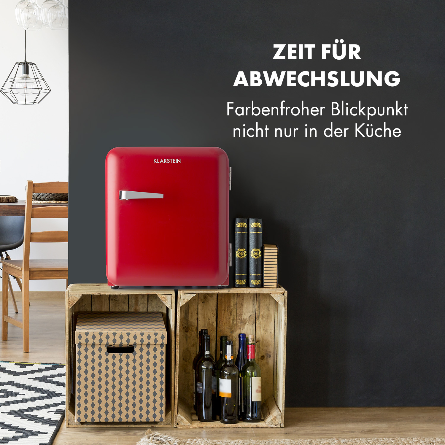 Un mini-frigo rosso su casse di legno, con bottiglie di vino e libri. Parete nera sullo sfondo.