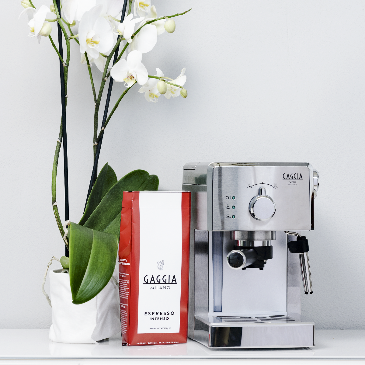 Máquina de espresso, bolsa de café roja y orquídea blanca sobre superficie blanca.